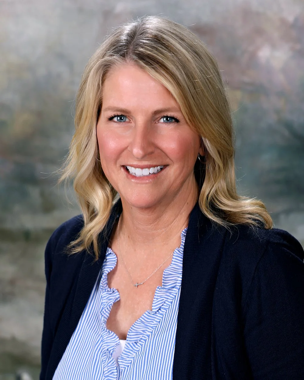A smiling blonde woman with wavy hair wearing a black blazer over a striped shirt, standing in front of a windowed background.