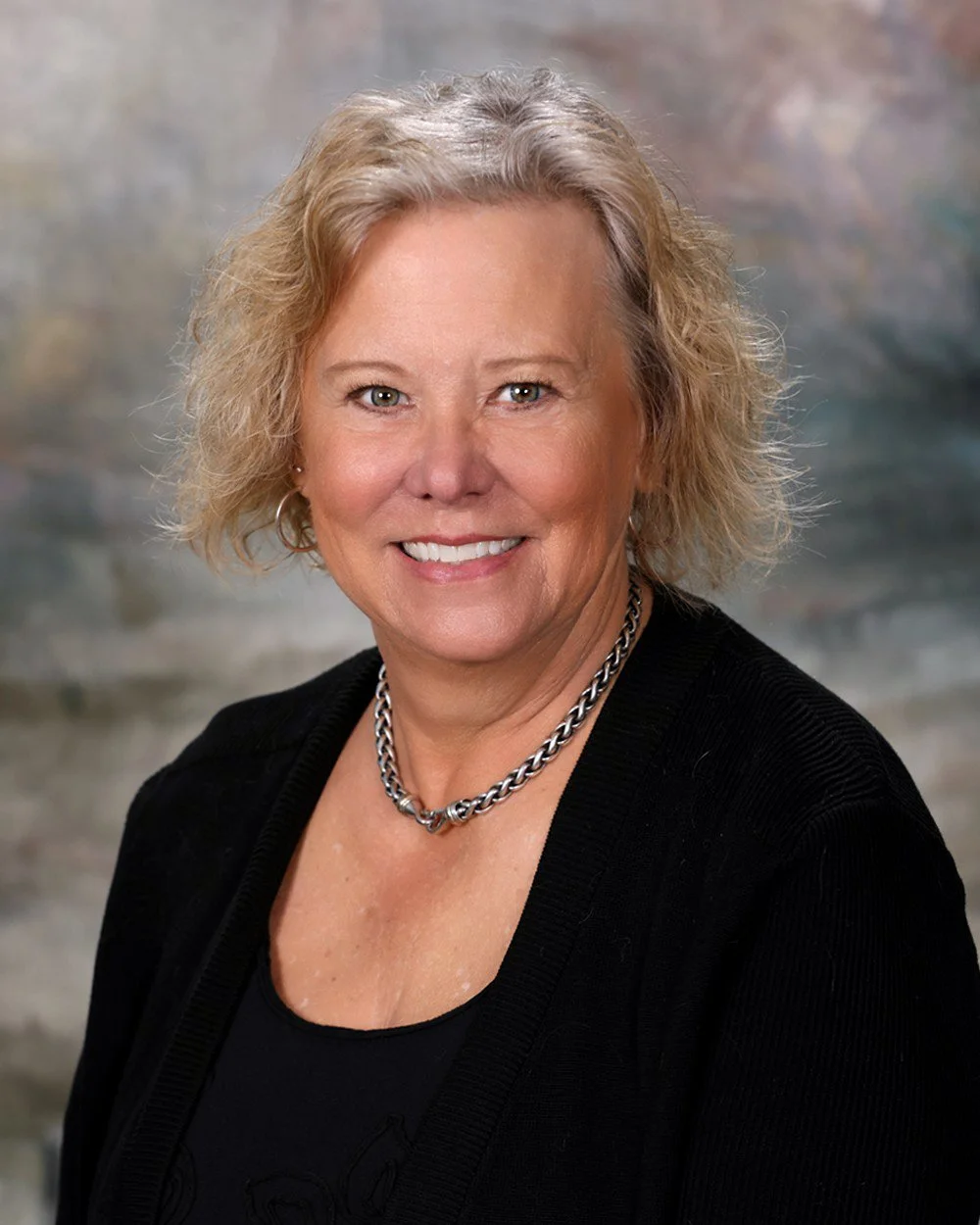A woman with curly blonde hair smiling, wearing a beige jacket and a gold necklace, standing in front of a light-colored wooden background.