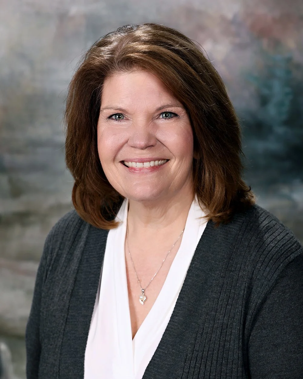 A woman with shoulder-length brown hair, blue eyes, and hoop earrings, smiling, wearing a white shirt and a gray cardigan with floral embroidery, standing in front of a rustic wooden window frame.