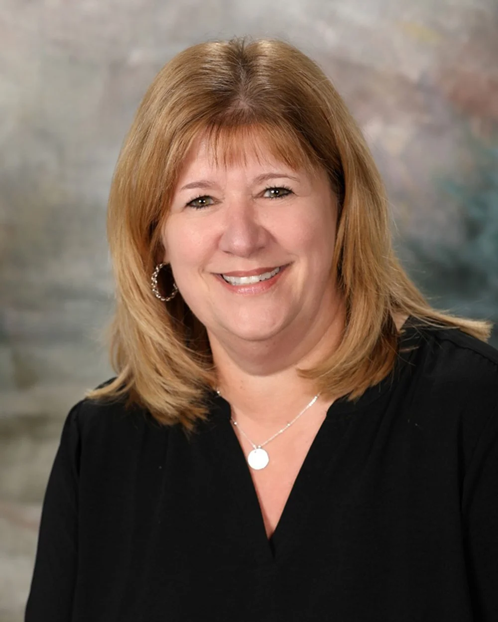 A smiling middle-aged woman with shoulder-length light brown hair, wearing pearl hoop earrings and a colorful floral blouse, standing in front of a rustic, whitewashed window frame backdrop.