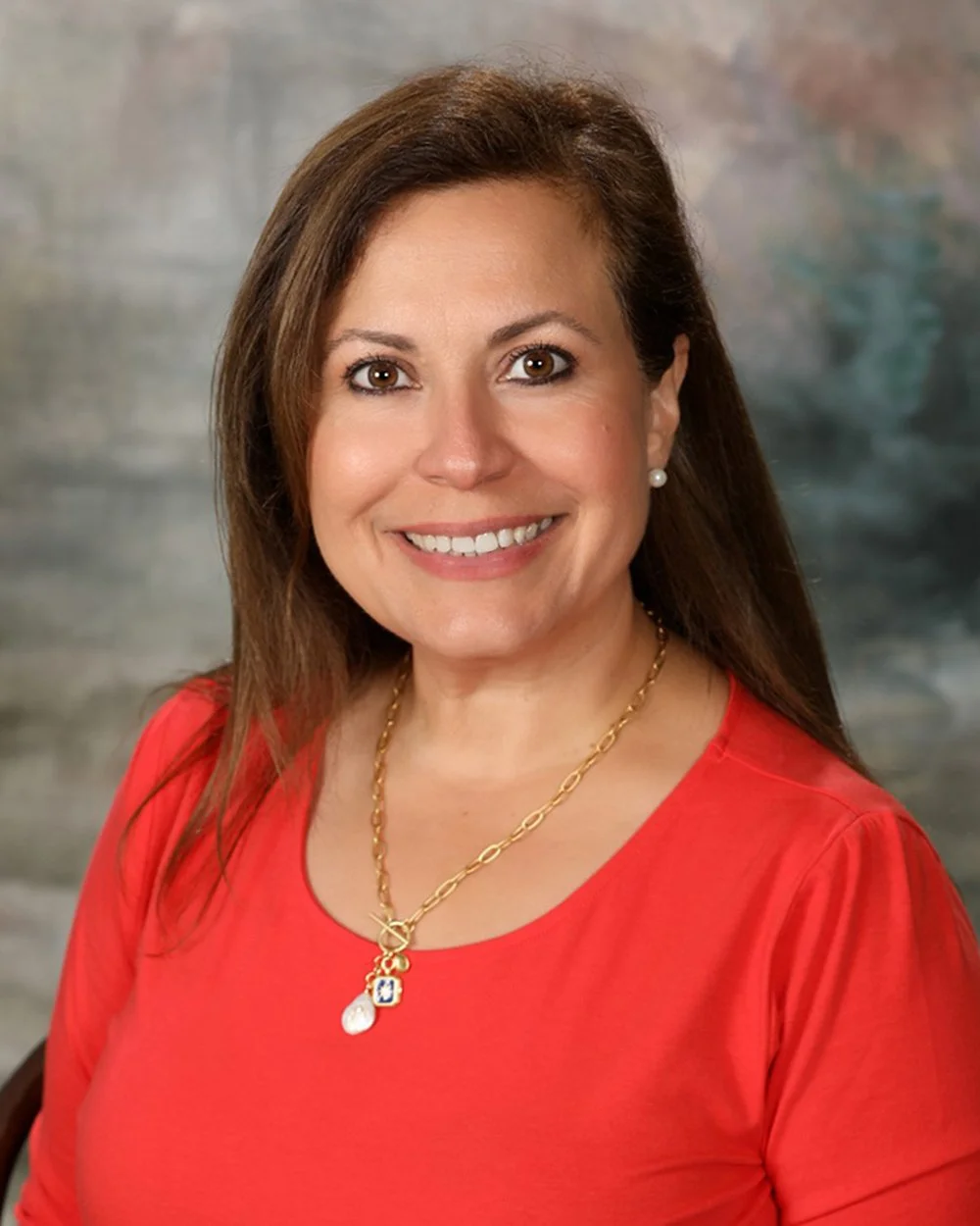 A woman with shoulder-length light brown hair, smiling, wearing a black and white striped shirt with a gold zipper, a pearl necklace and pearl earrings, standing in front of a background with a grid of white wooden panels.