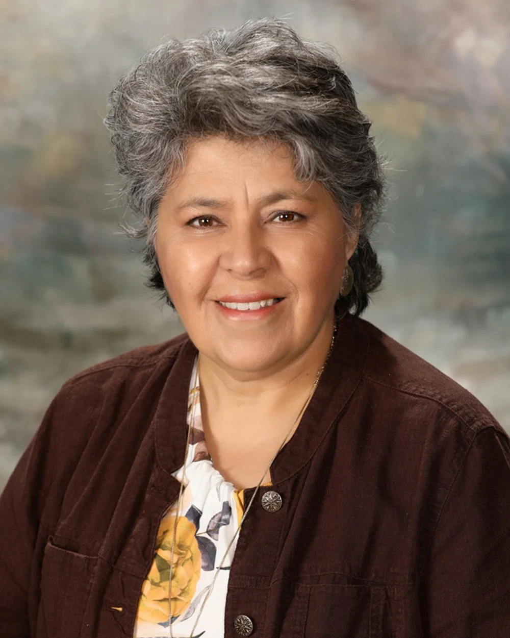 A smiling woman with short, curly gray hair wearing a yellow jacket over a black top, accessorized with a silver necklace and earrings, standing in front of a background with white window panes.
