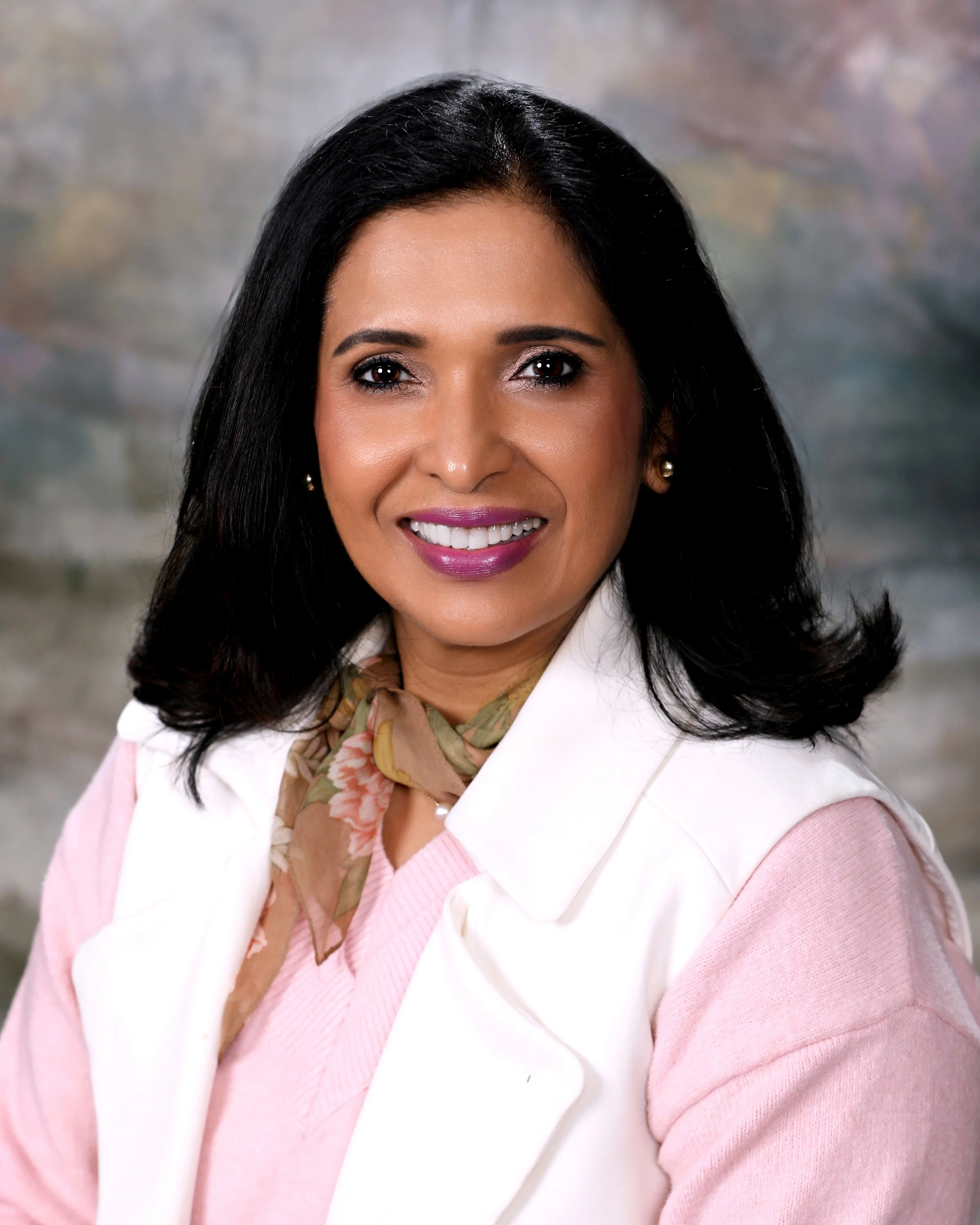 A woman with long black hair, wearing pearl earrings and a pearl necklace, smiles at the camera in front of a light-colored background with a window grid pattern.