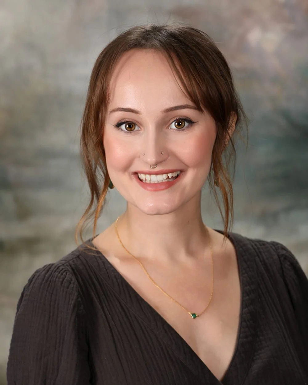 A young woman with short, wavy brown hair smiling at the camera, wearing hoop earrings and a green top, standing in front of a white wooden window frame background.