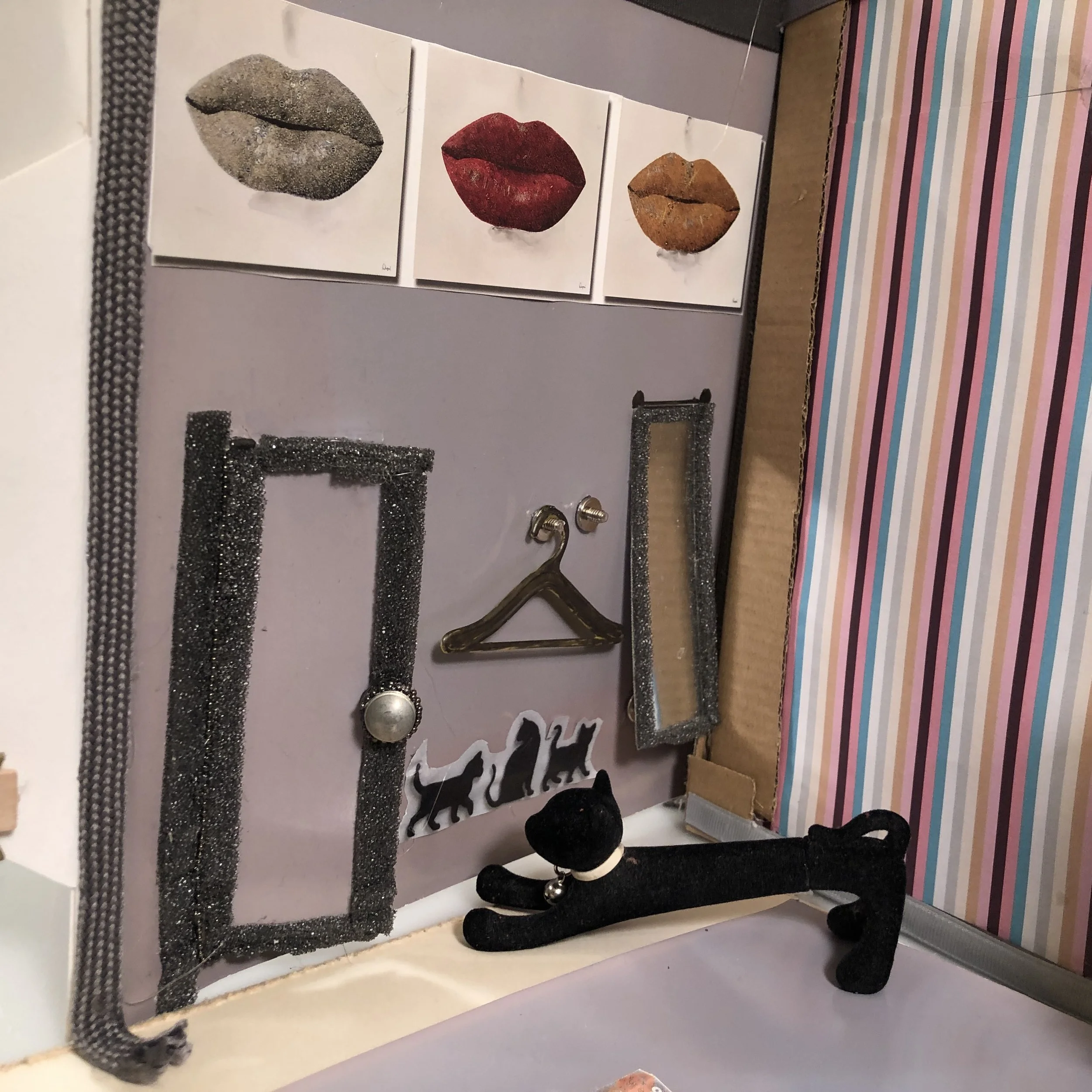A display board with three canvas prints of lips, a small black plush cat, and a black plush dog, with a striped background on the right side.