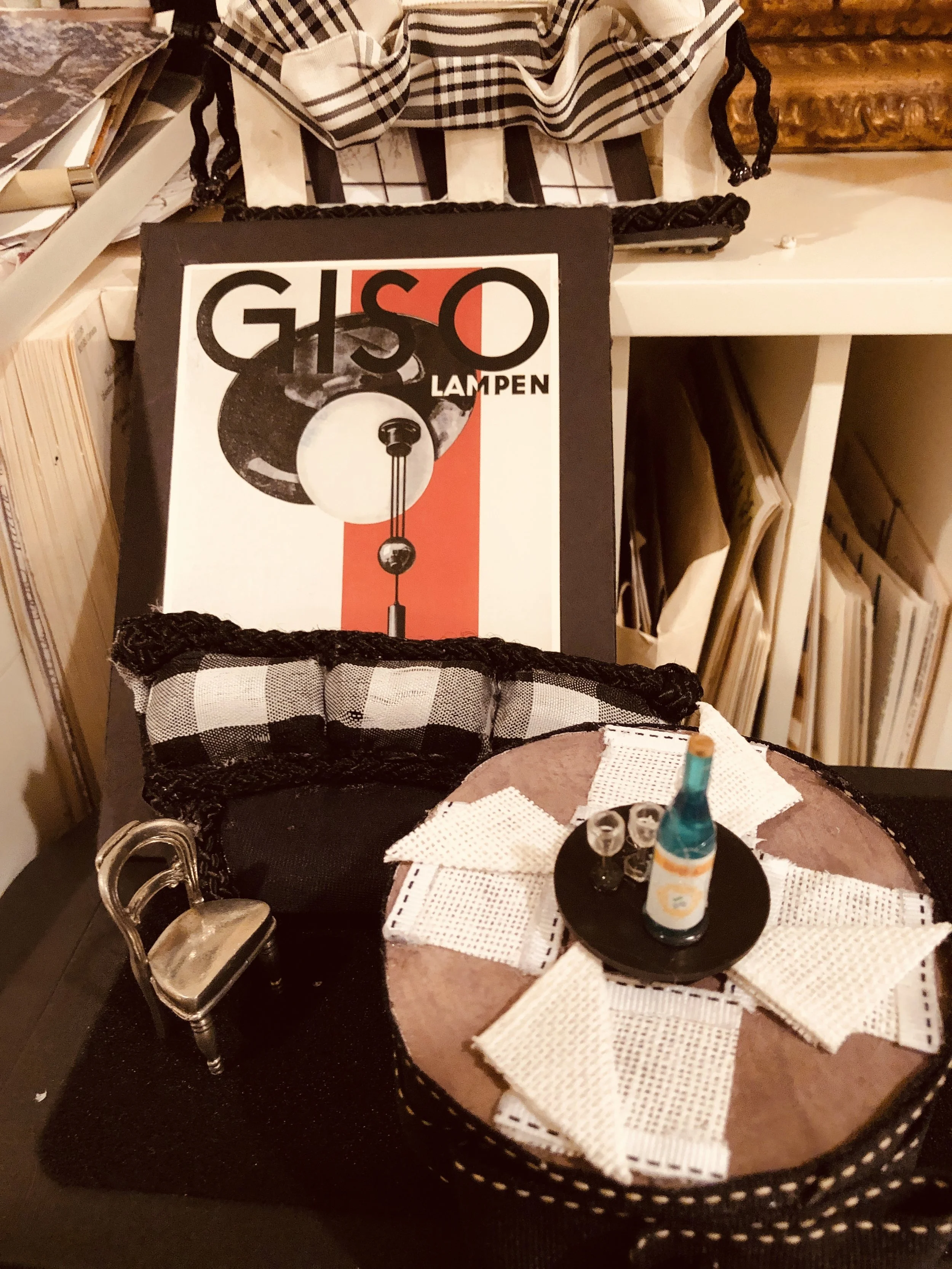Here, small round table with a miniature bottle, two miniature glasses, and white cloth napkins. In the background, a vintage black and white poster of a Giso lamp, a black-and-white checkered cloth, and a bookshelf filled with books and magazines.