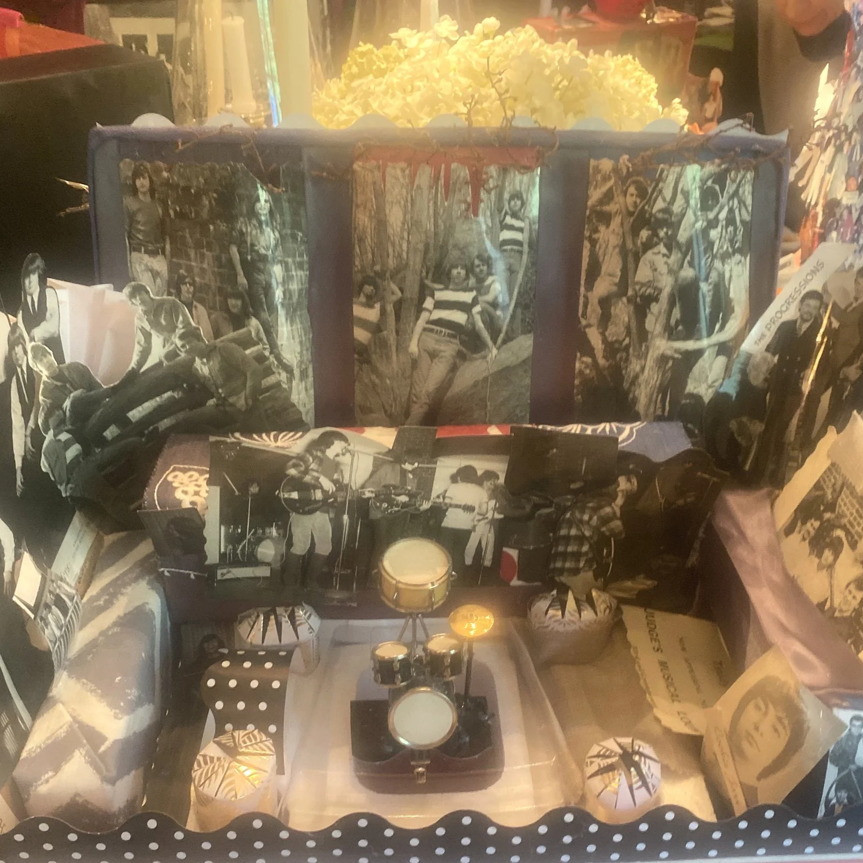 A display featuring black and white photographs of a young woman, a mini drum set, candles, and various decorative items, with a yellow flower arrangement in the background.