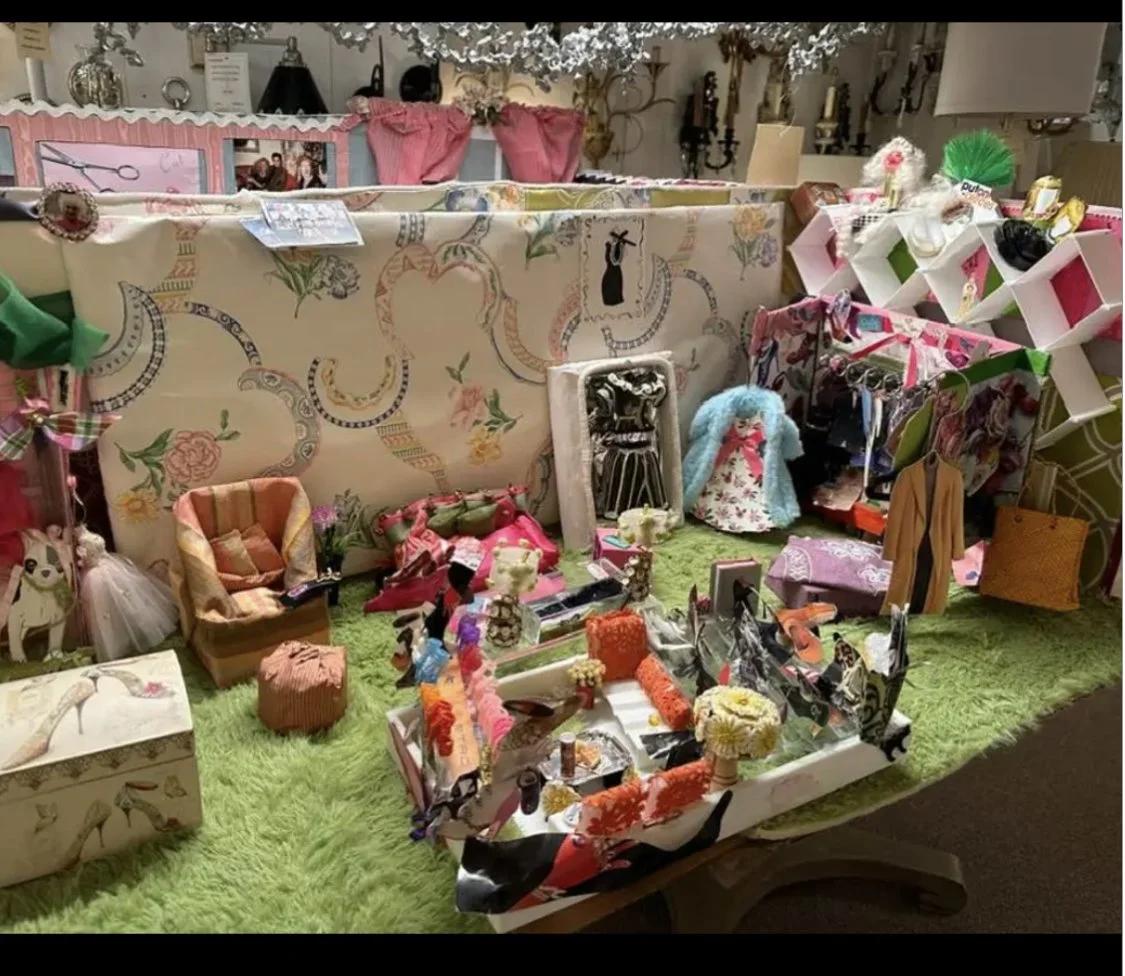 Display of various miniature home decor and fashion items, including clothing, accessories, and small furniture pieces, arranged on a green fuzzy surface with a decorative background.