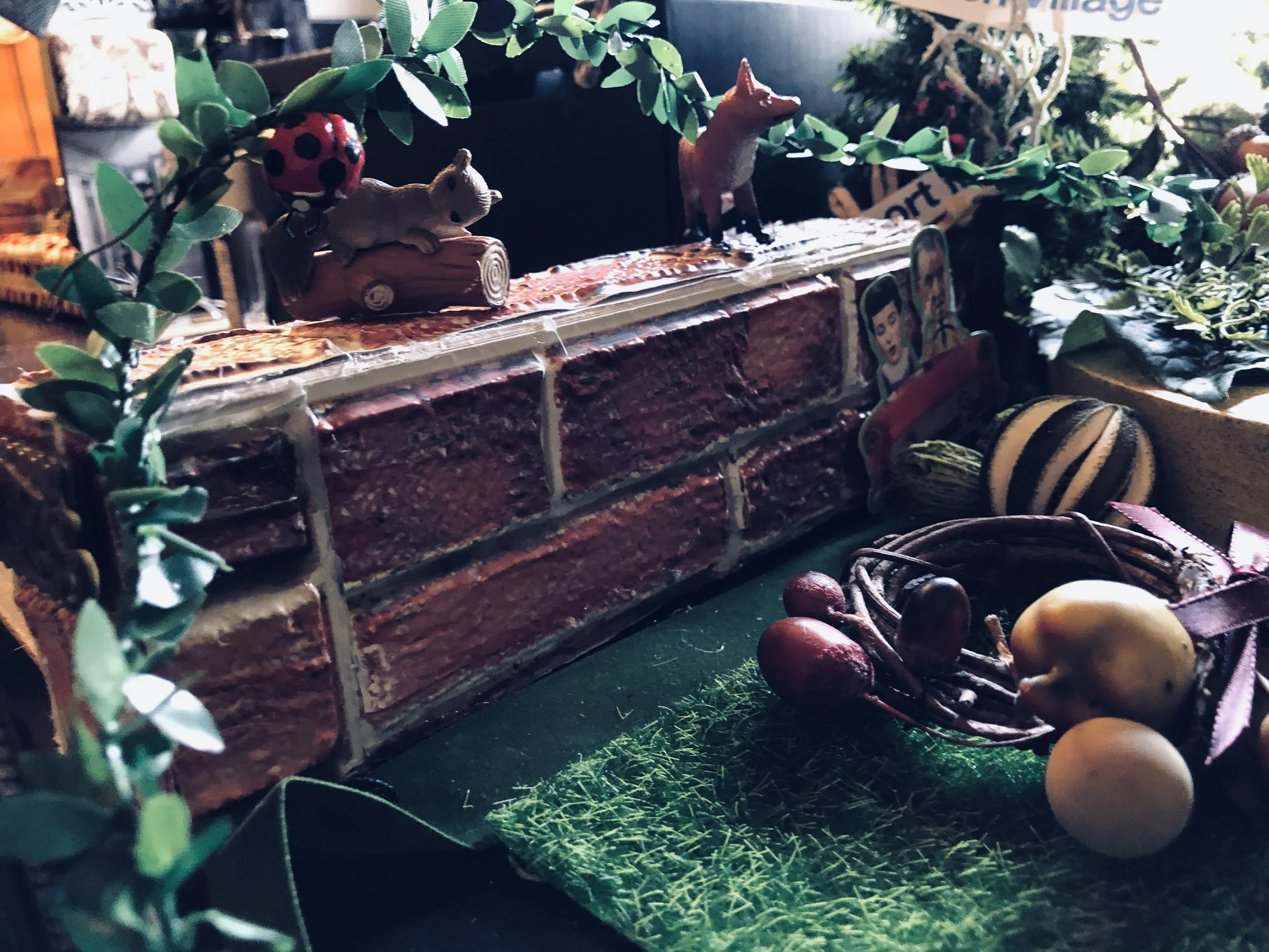 Decorative scene with small animal figurines, artificial green foliage, a brick-patterned surface, and a bunch of miniature vegetables including pears and grapes, arranged on a table with a Christmas or festive theme.