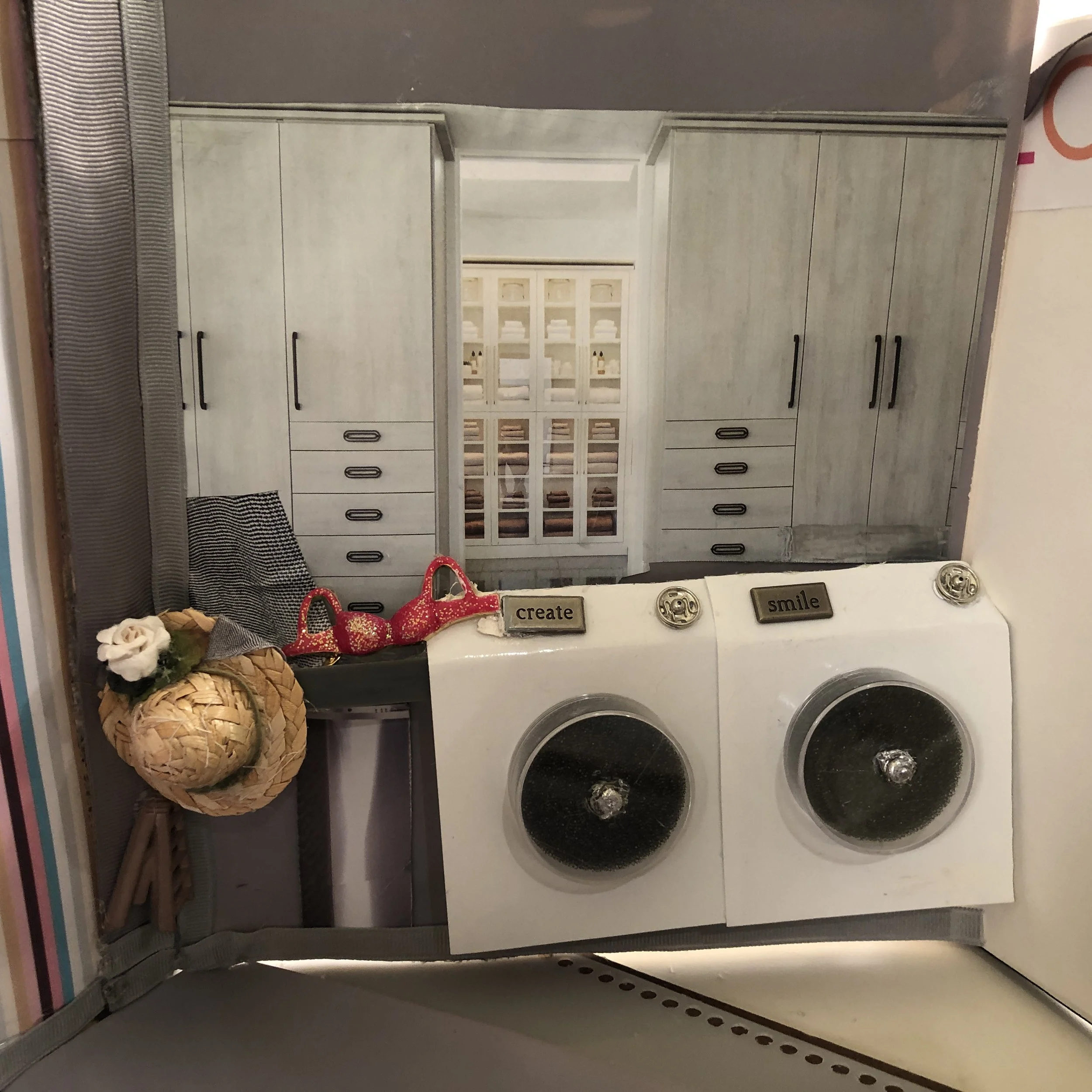 A miniature laundry room scene with two front-loading washing machines. There are decorative motivational signs reading 'create' and 'smile' on top of the washing machines. A small woven basket with a white flower and greenery hangs on the left side.