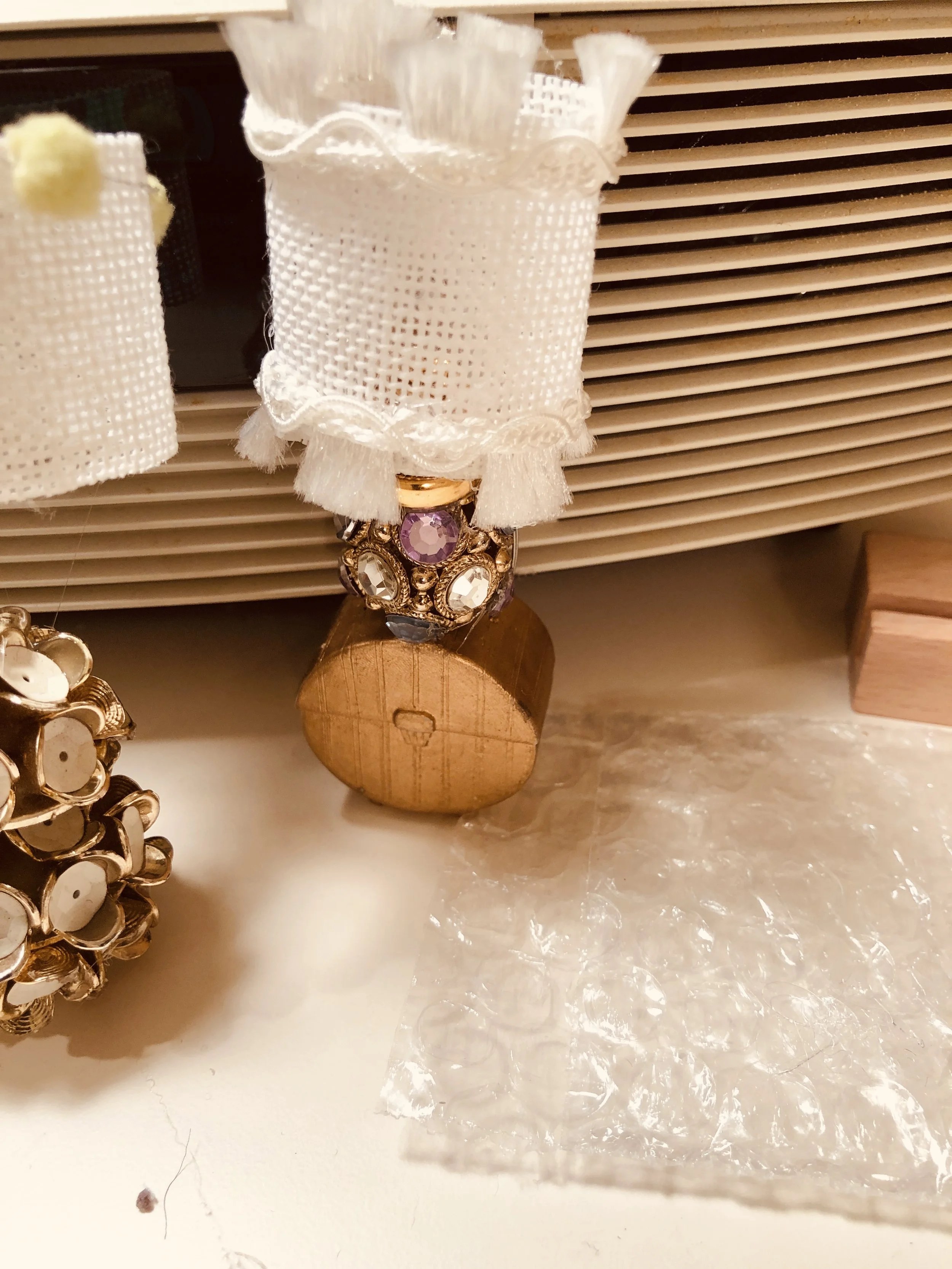 Close-up of a decorative lamp with a white fabric shade and embellishments, sitting on a wooden base, placed on a textured surface next to other jewelry or decorative items.