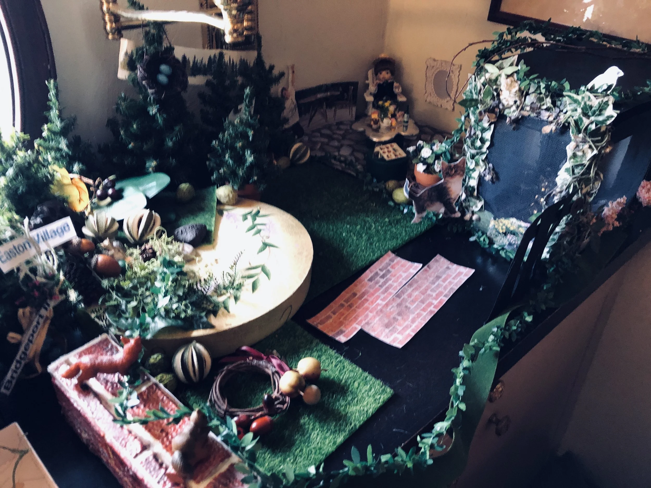 A miniature Christmas scene with small decorated trees, stuffed animals, and faux brick and grass decorations, set on a table with a green surface and surrounded by Christmas greenery.