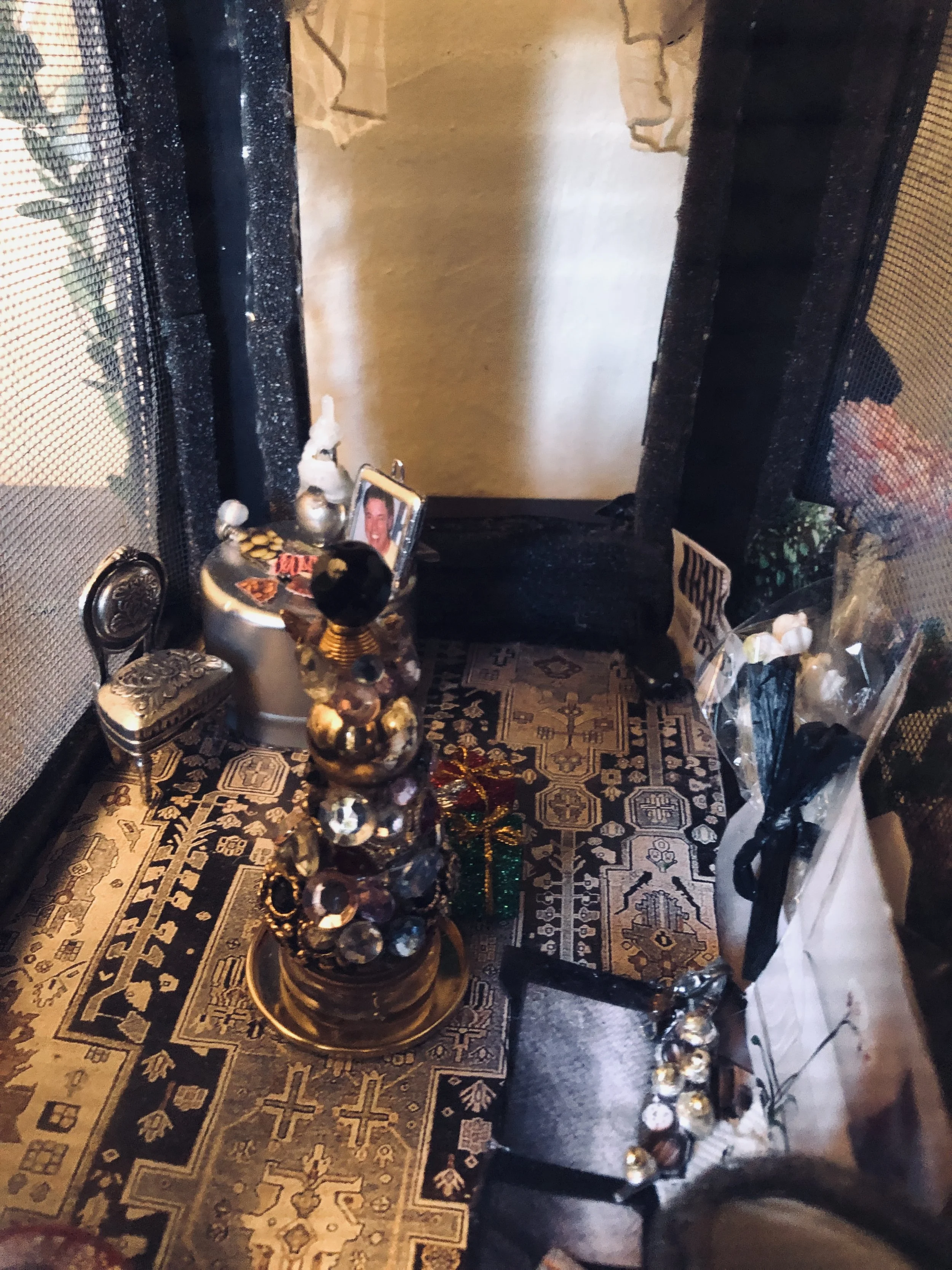 A small table with a black and gold patterned cloth, decorated for the holidays. Items on the table include a frame with a photo of a man, a small silver chair, a decorative teapot, a miniature Christmas gift, several beads or ornaments, and a plasti