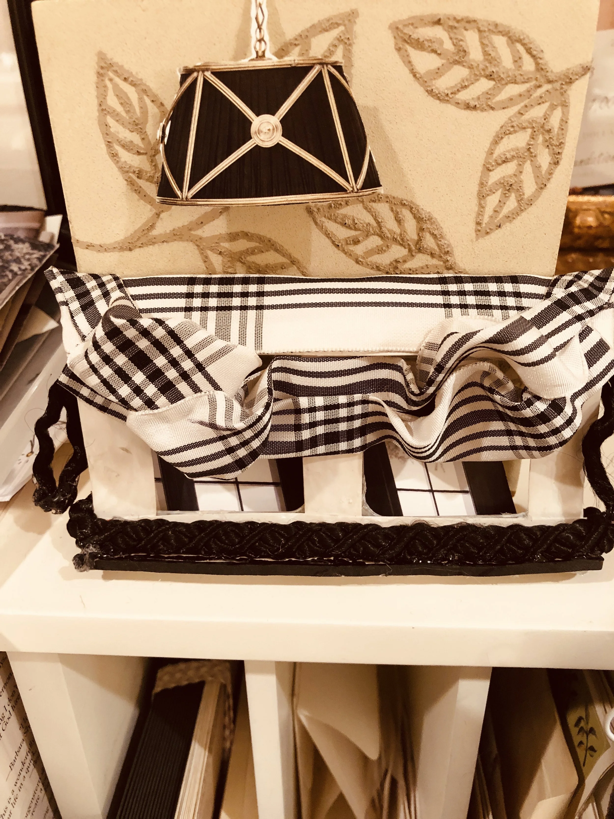 A black and white checkered fabric or napkin in a black basket with braided edges, placed on a white shelf with other items underneath. In the background, there is a decorative wall with a black lampshade and a leaf pattern.