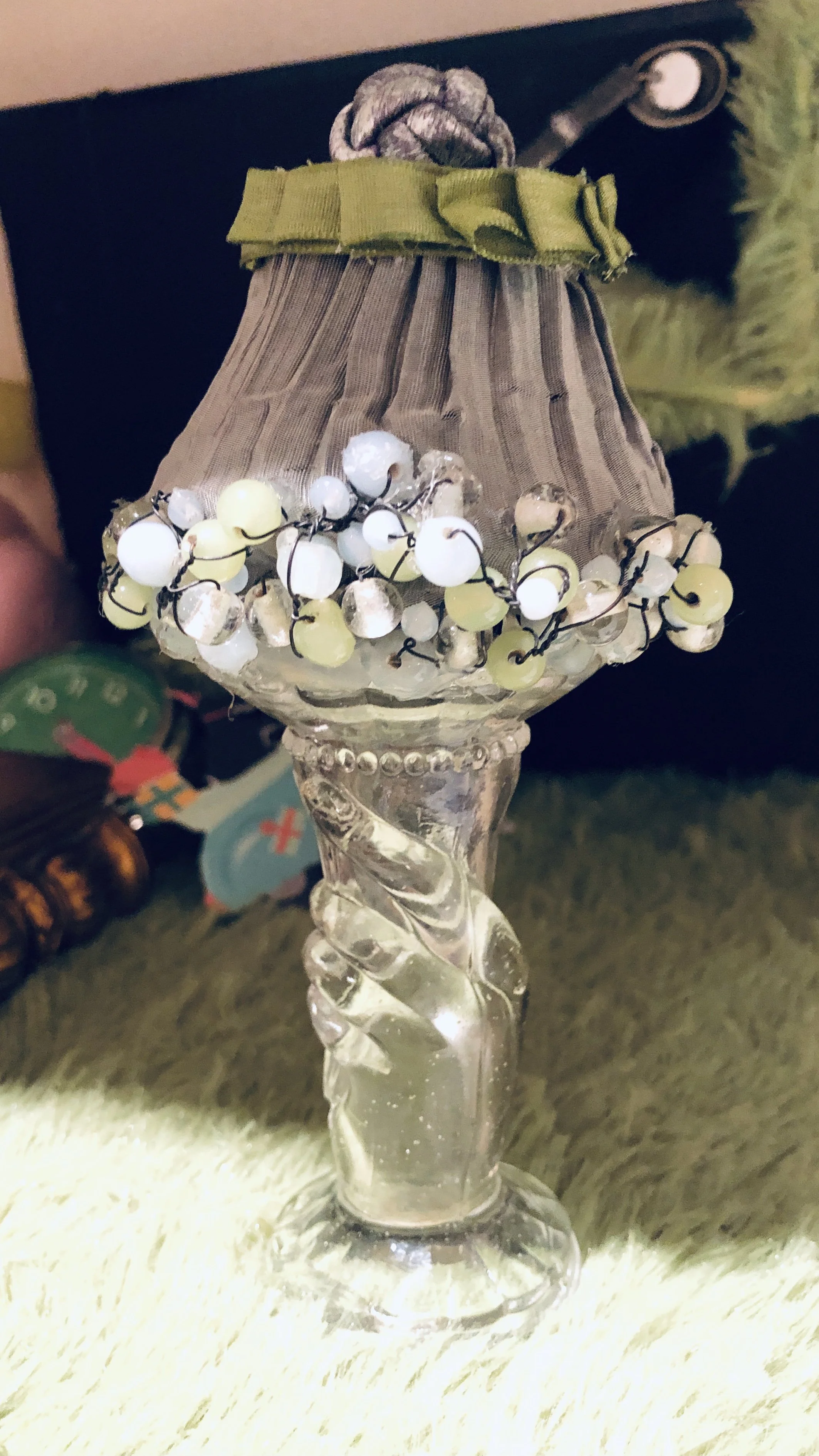A decorative glass lamp with a twisted glass hand holding the lampshade, which is wrapped with a black fabric and adorned with a beaded jewelry around the edge.