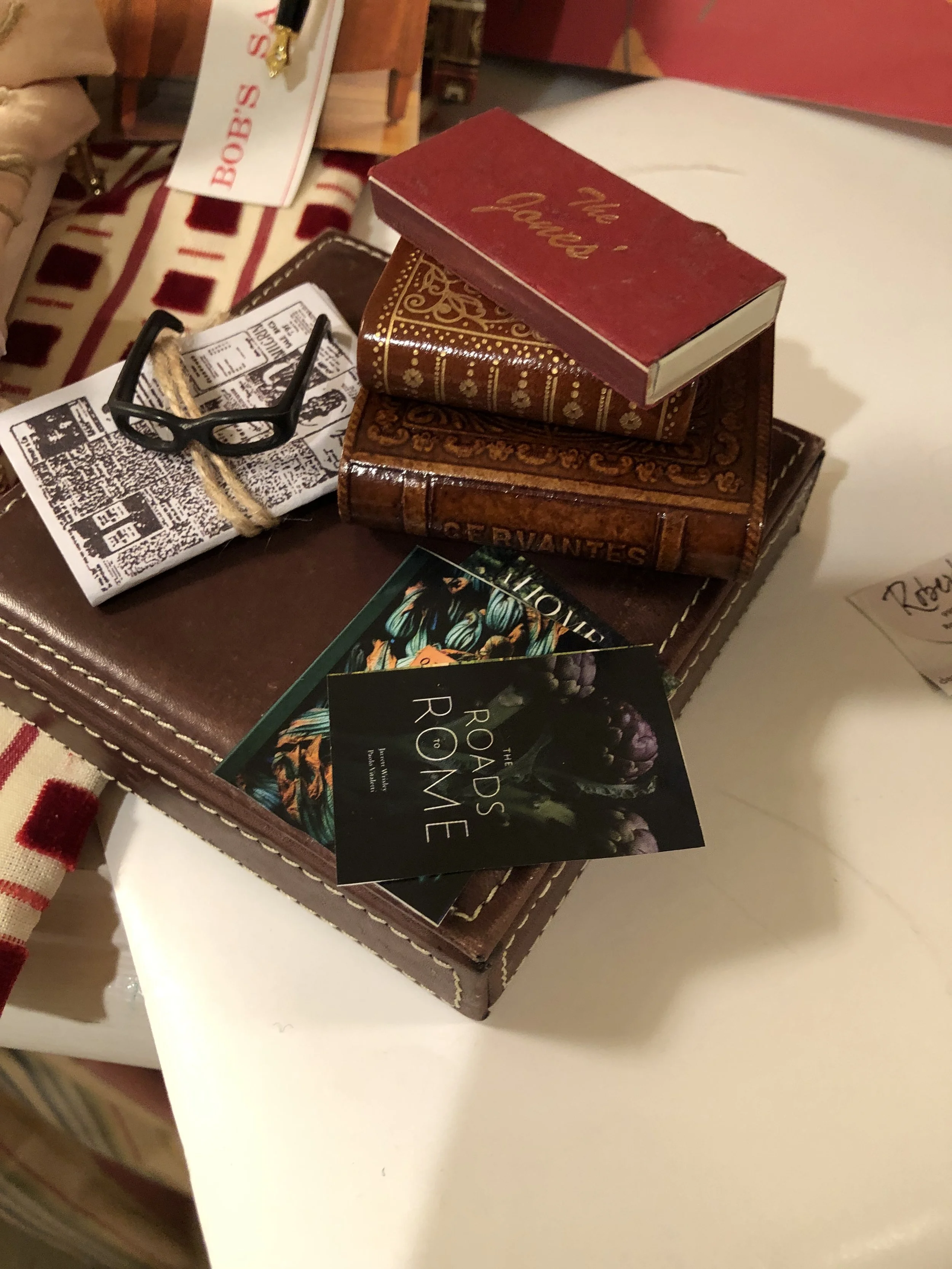 Repurposed and assembled Miniature Stack of vintage books, a pair of black glasses, and printed promotional cards on top of a brown leather wallet or cover.
