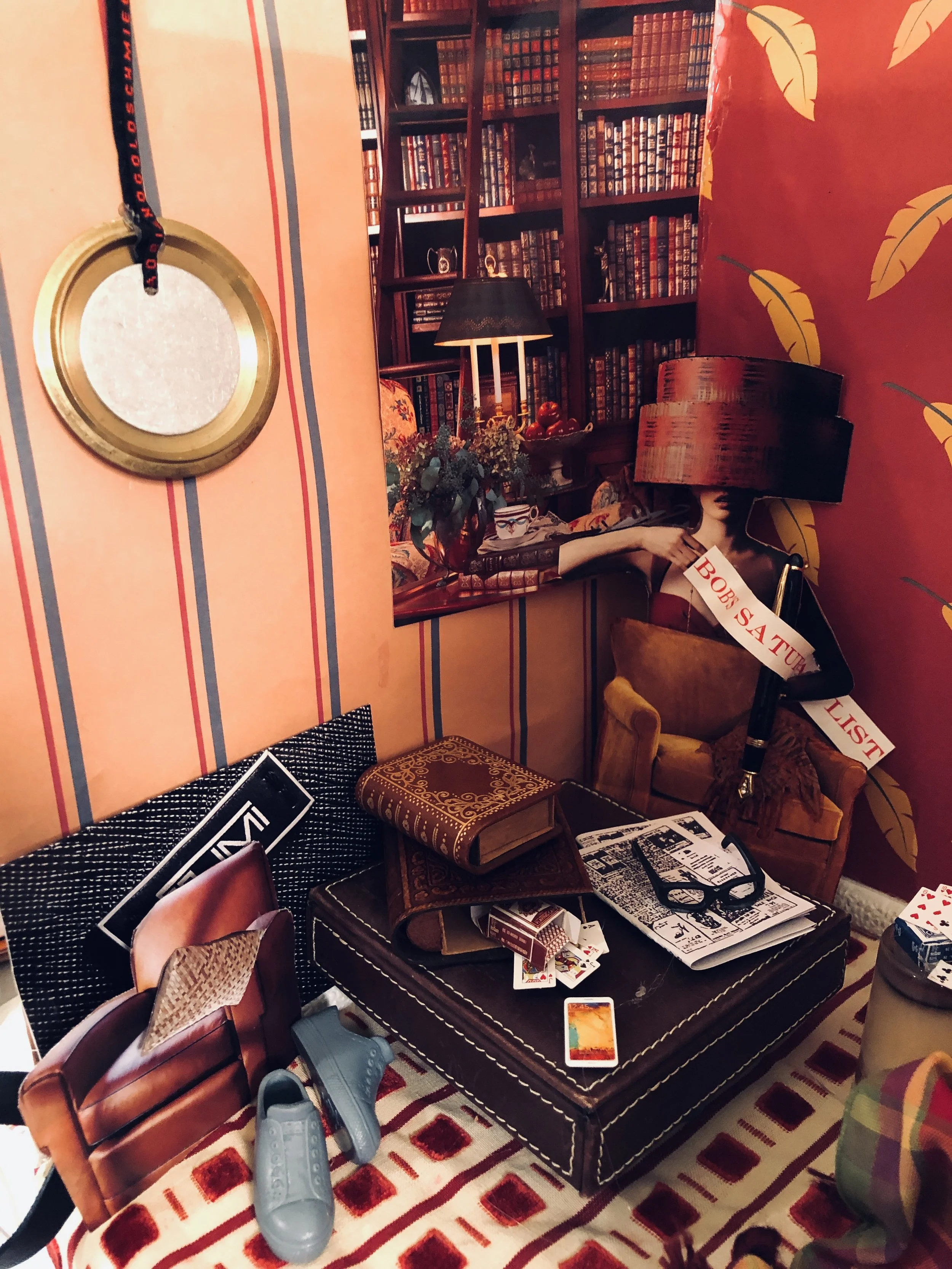 Eclectic living room corner with a dark leather ottoman holding books, magazines, glasses, and a phone, surrounded by colorful decor including a red and yellow wallpaper, a shoe, and a poster of a woman with a lampshade over her head, with a bookshel