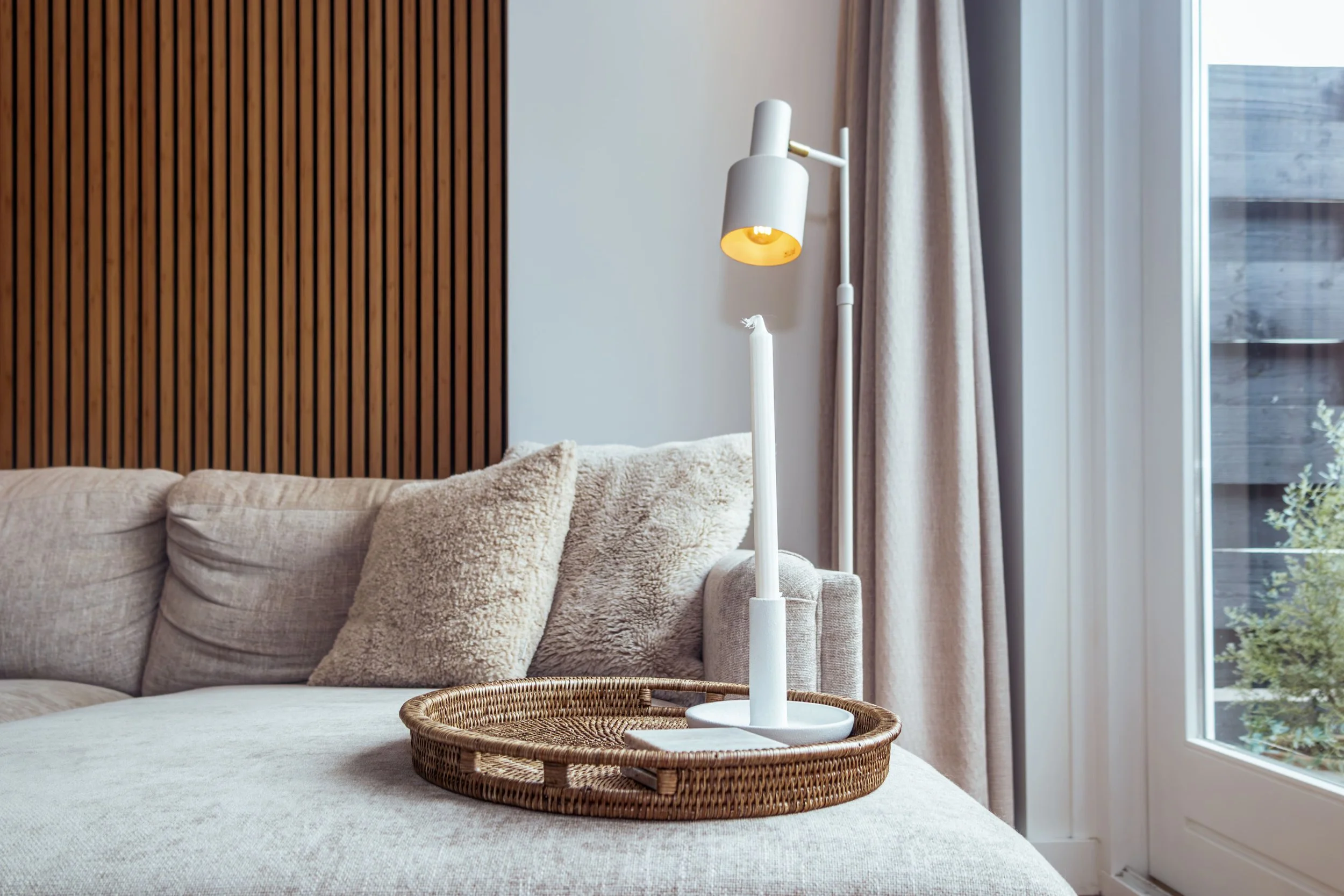 Woonkamer met beige bank, kussens, houten wand, staande lamp en grote raam met beige gordijnen.