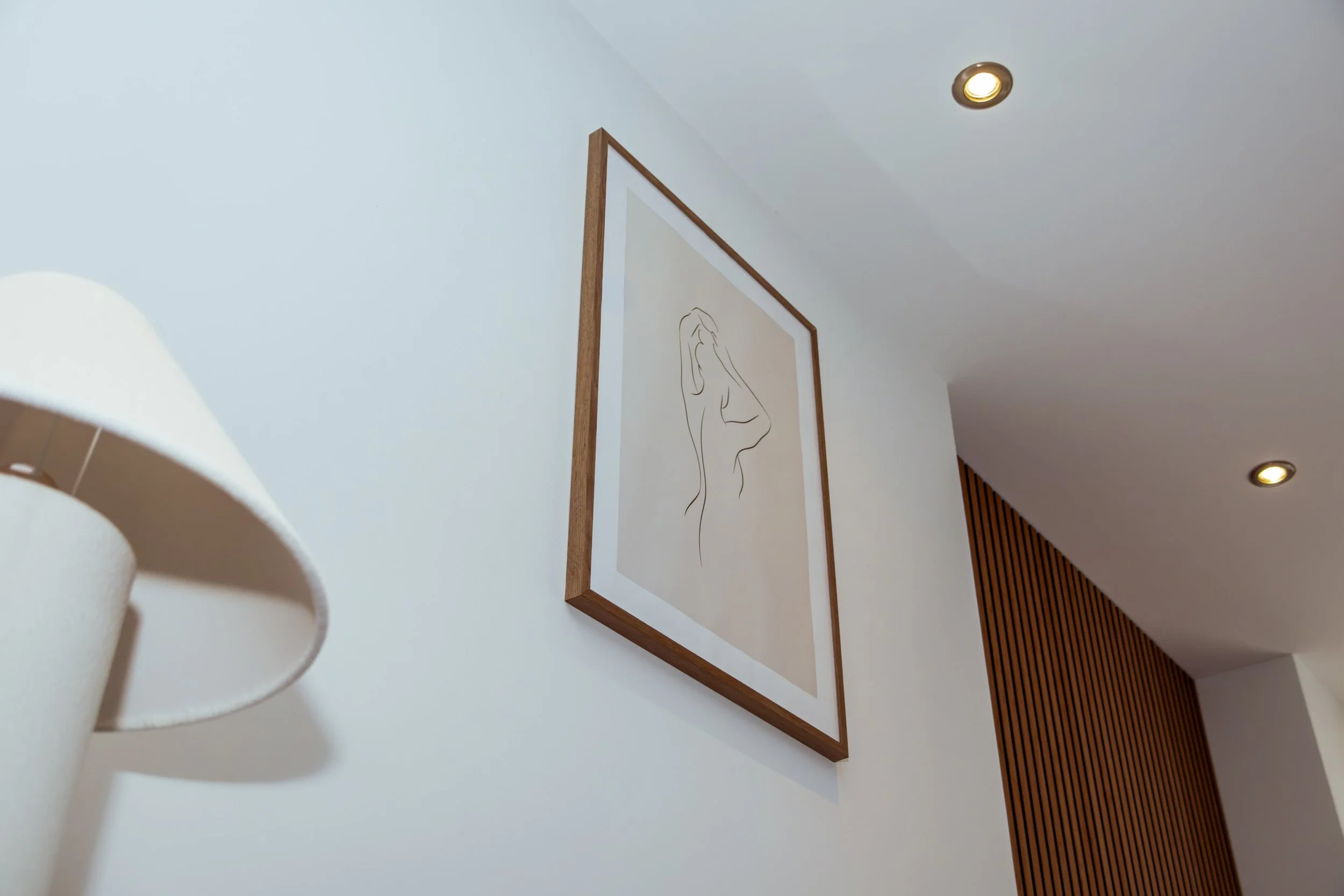 Een wit geschilderde muur met een kader met een lijntekening van een gezicht in minimale stijl en twee ingebouwde plafondlampen.
