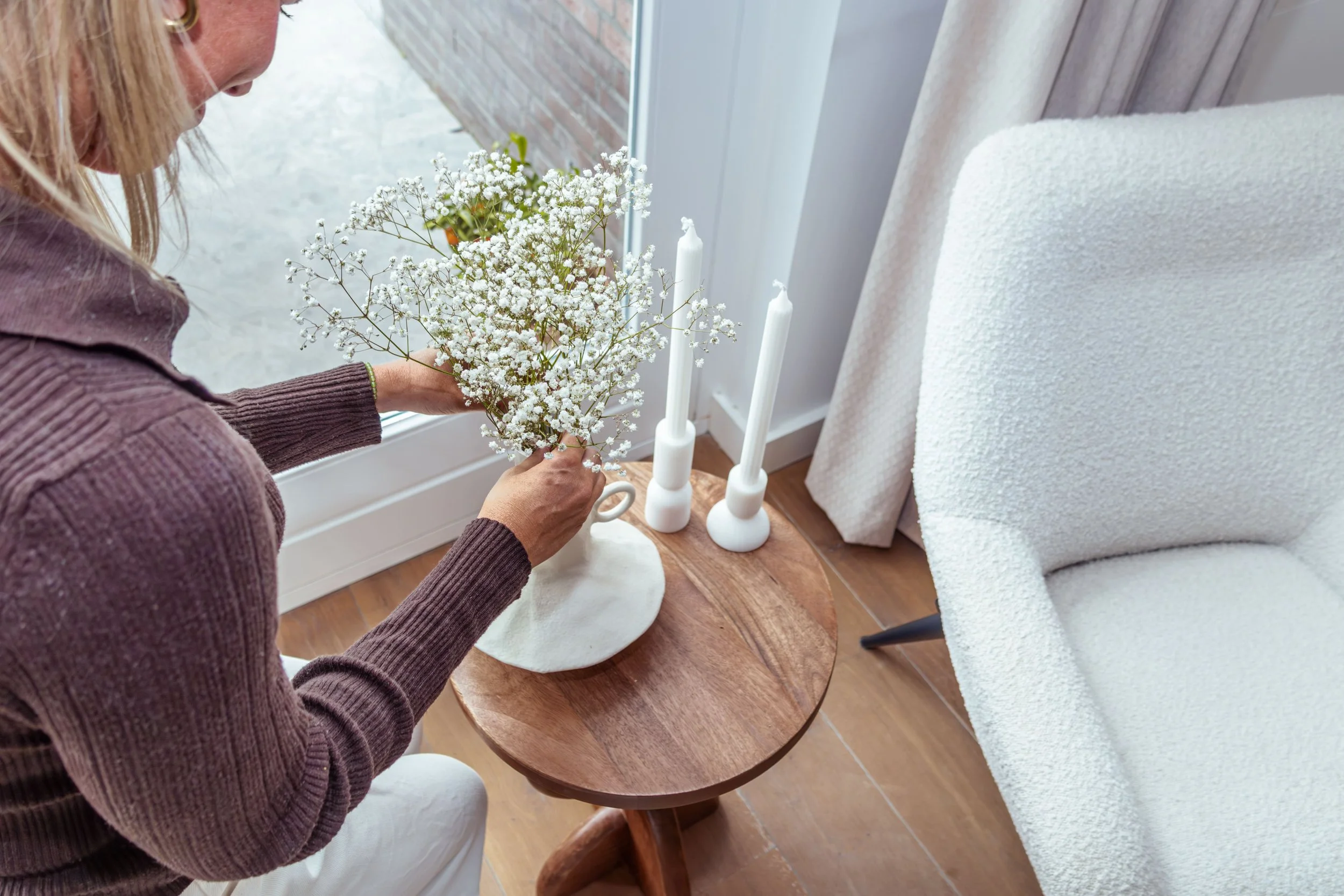 Vrouw die witte bloemen plukt naast twee witte kandelaars op een houten tafel naast een raam en een witte stoel.
