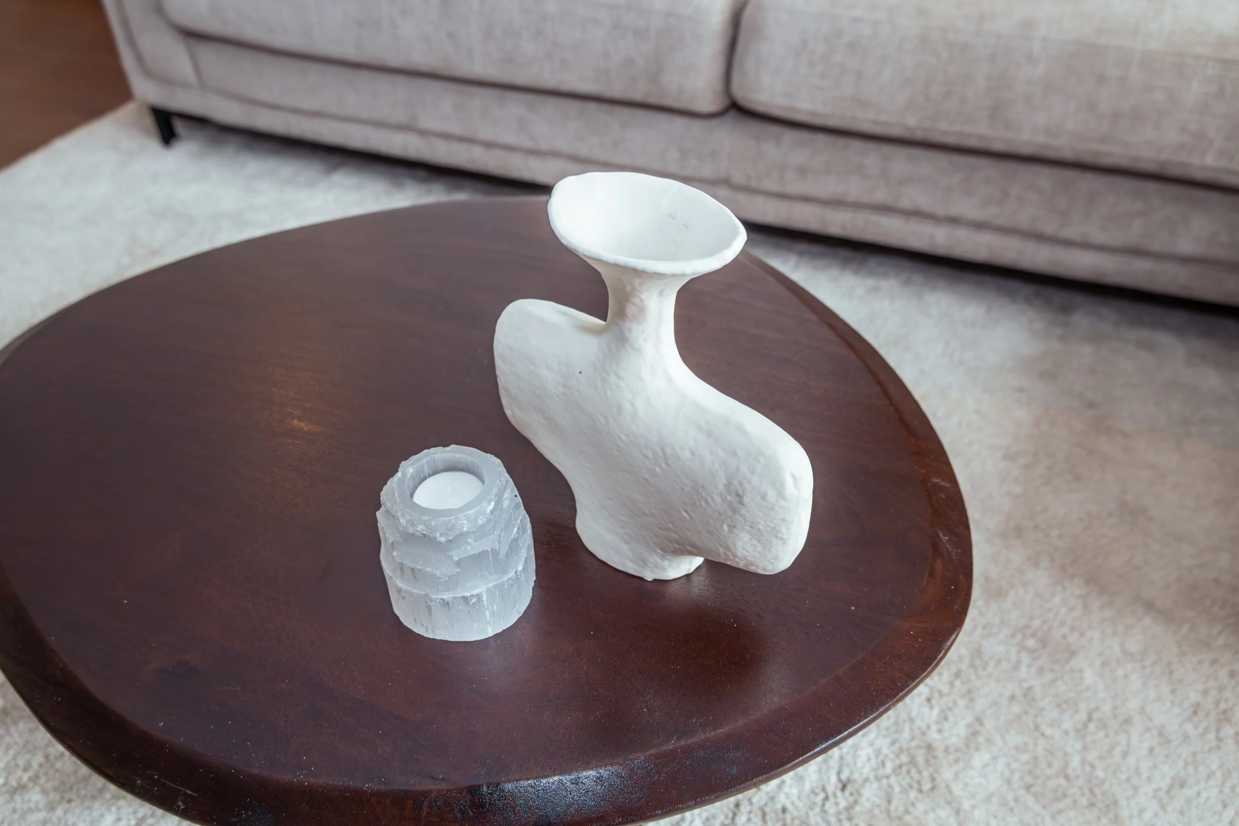 Wit decoratief object op een ronde houten tafel met een kaars in een glazen houder, in een woonkamer met beige tapijt en een bank op de achtergrond.