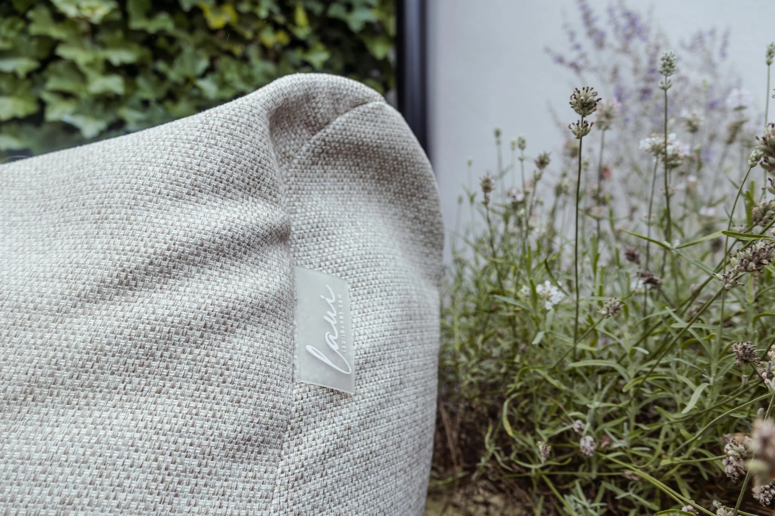 Een lichtgrijze stoffen tas met een label, liggen naast bloemen en planten in een tuin.