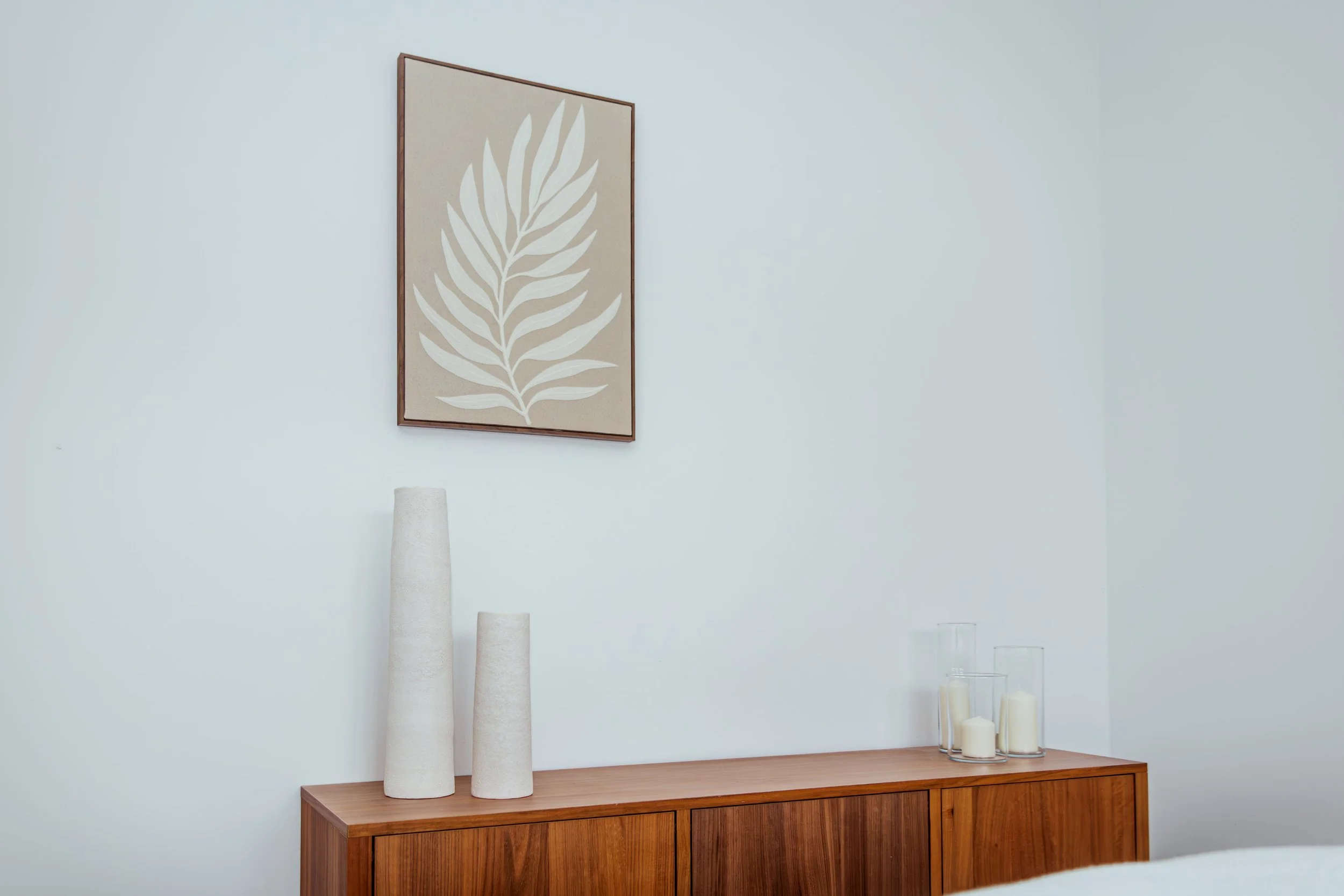 Een houten dressoir met witte vazen en kaarsen, hangend aan een witte muur met een schilderij van een blad.