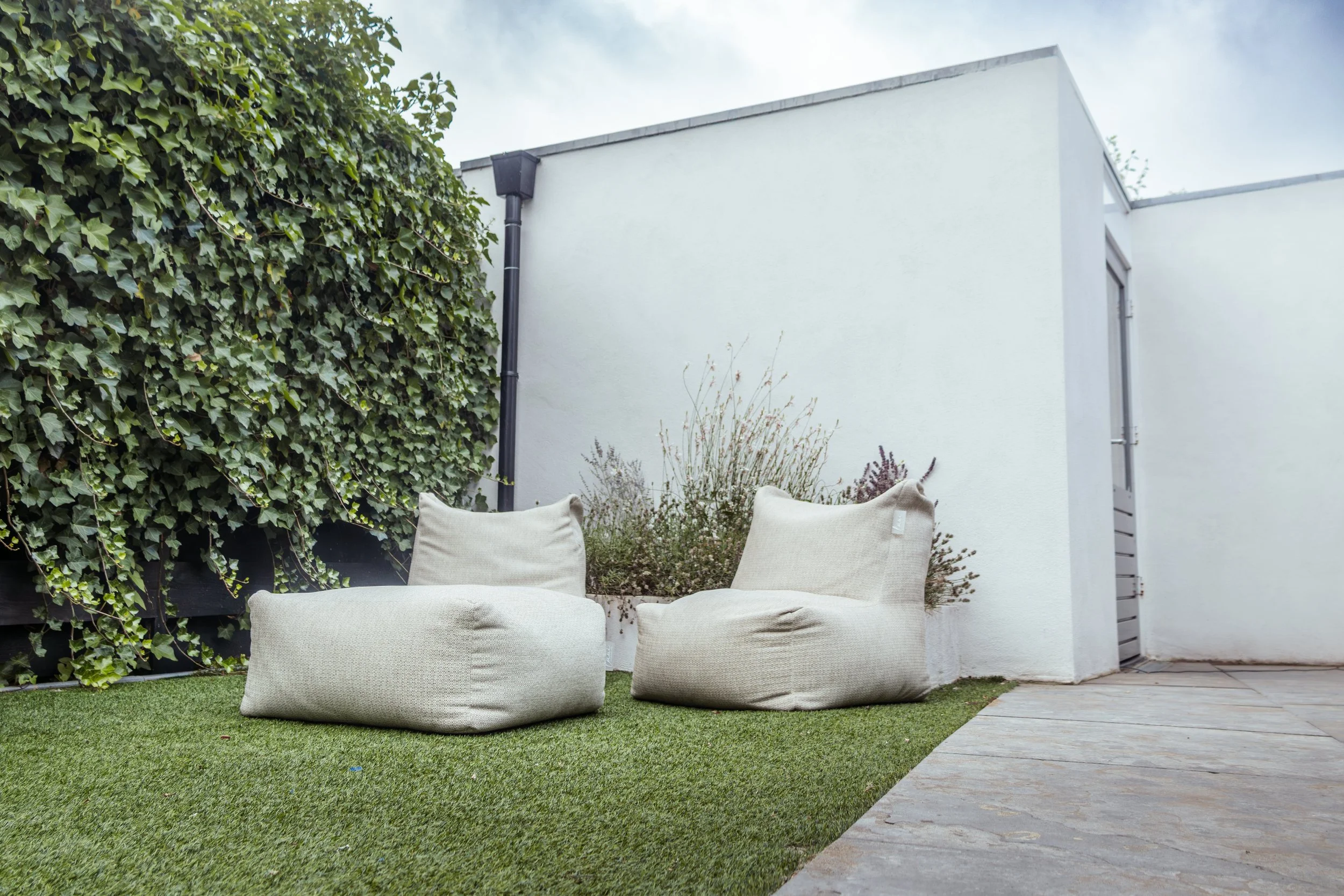 Twee beige zitzakken op kunstgras in een kleine tuin met witte muur en groene heg.