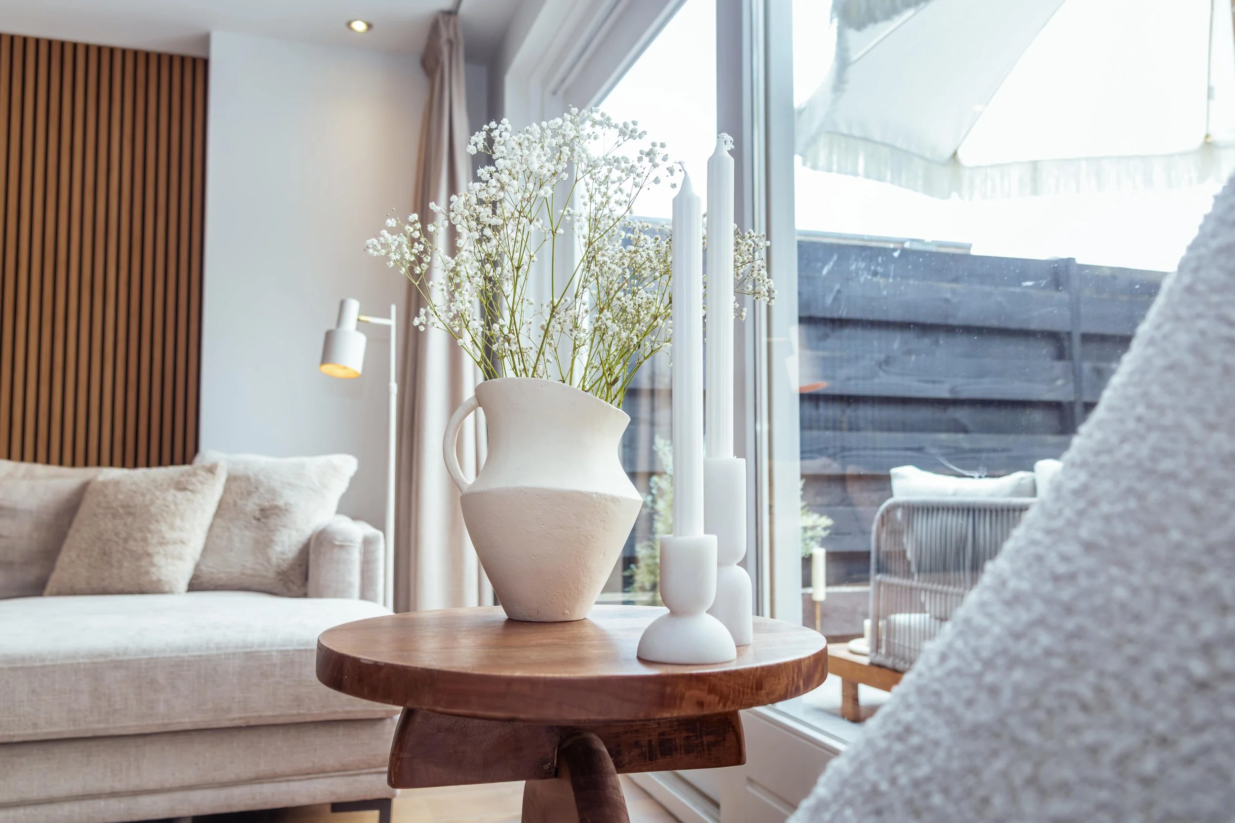 Binnenkamer met witte bank, houten salontafel met witte vaas met bloemen en witte kandelaars, grote ramen met uitzicht op een terras met parasol.
