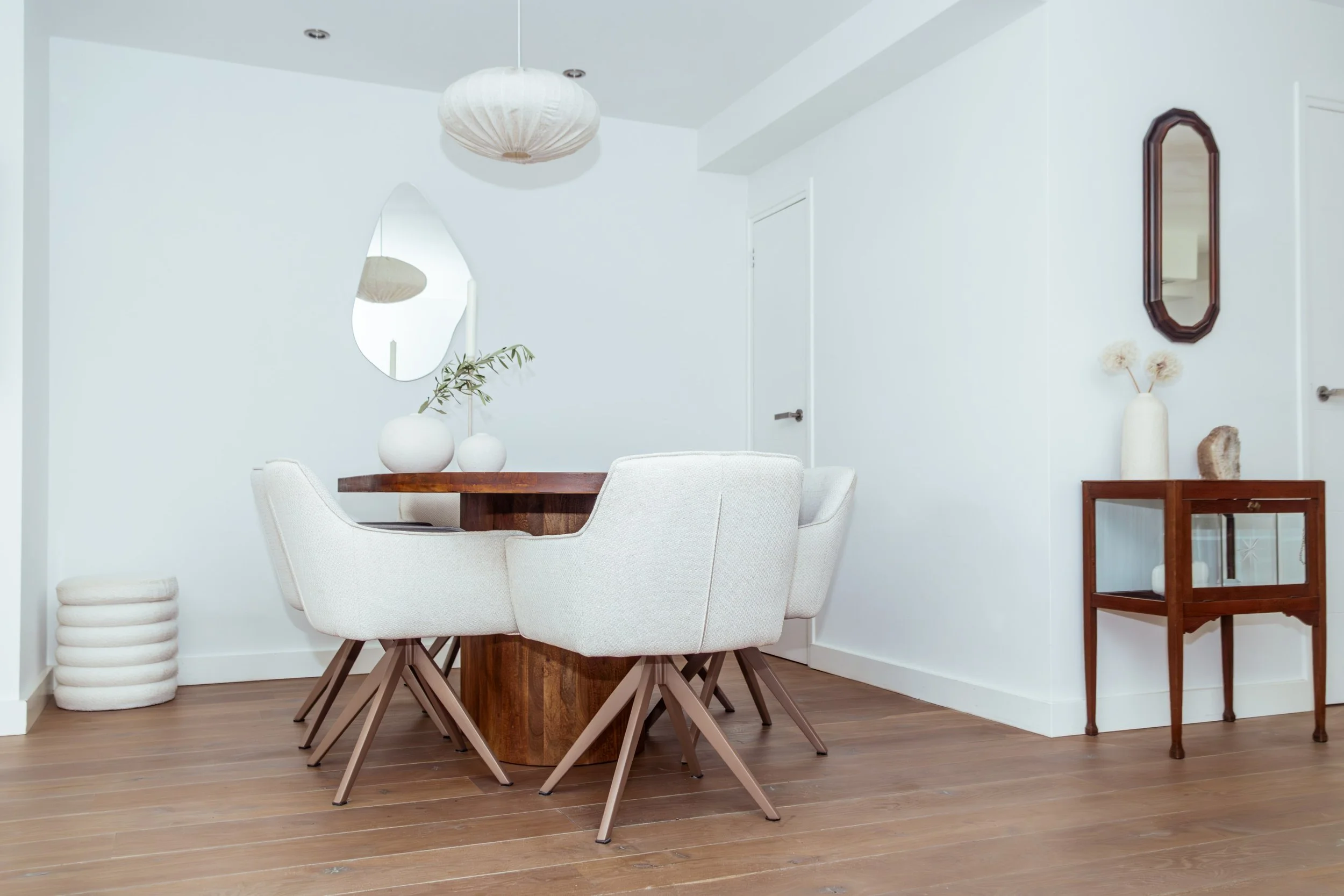 Woonkamer met witte stoelen, houten tafel, wit vazi en spiegel op witte muur, houten vloer, houten bijzettafel met vaas en decoratie.