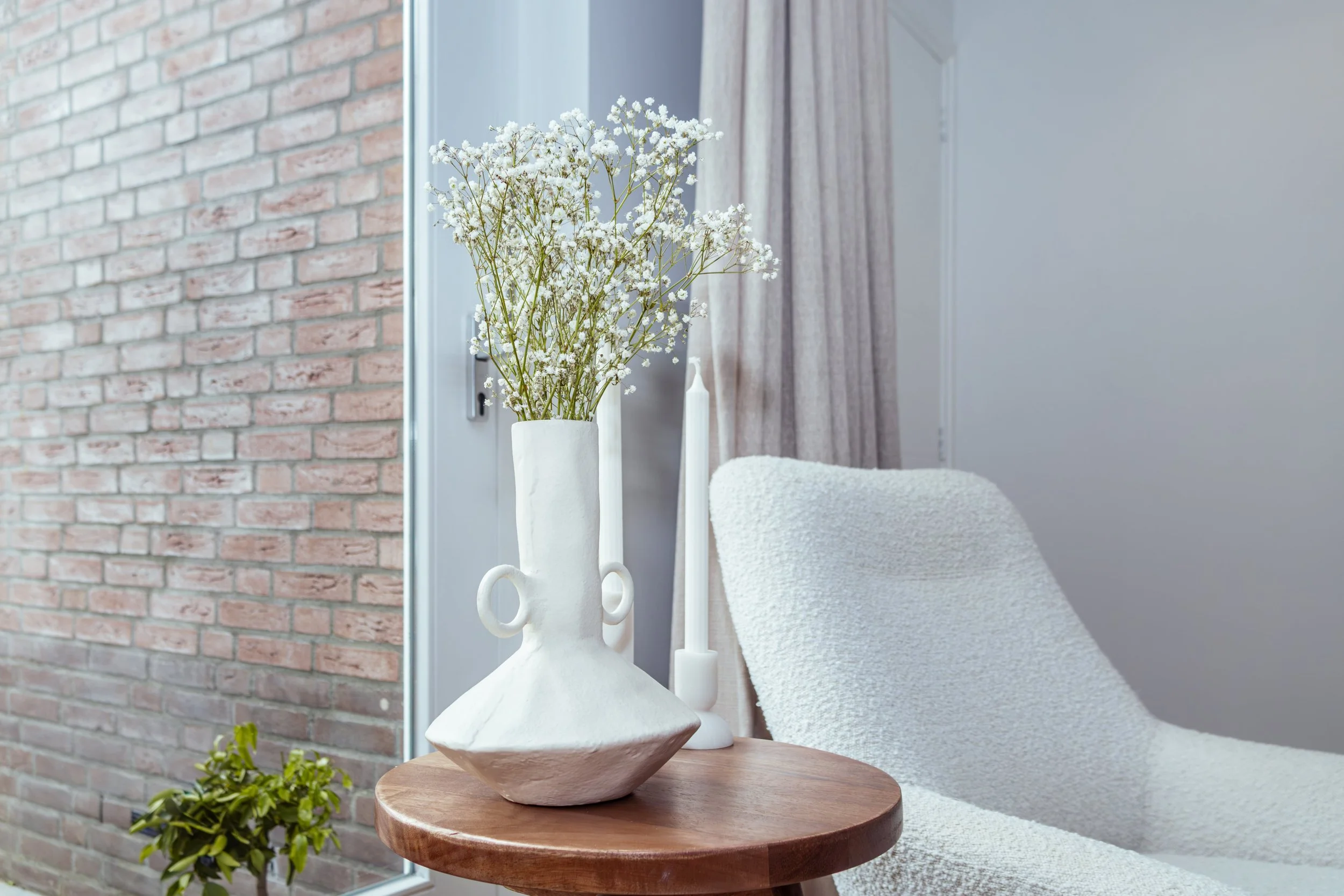 Binnenhoek met een ronde houten tafel, witte vaas met witte bloemen, witte kaars, witte stoel met textured bekleding, een raam met beige gordijnen, een bakstenen muur en een groene plant.