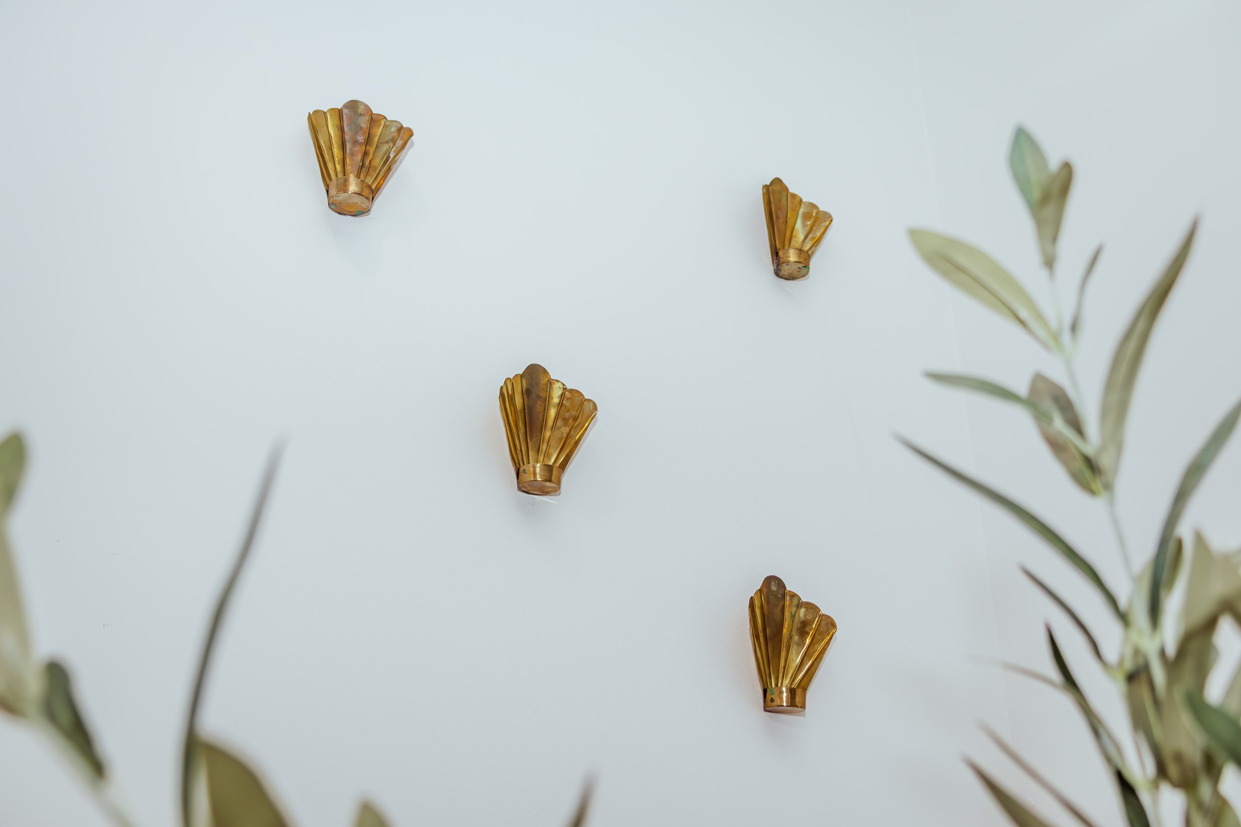 Vijf gouden wandhaken in een patroon op een witte muur, omringd door groene bladeren.