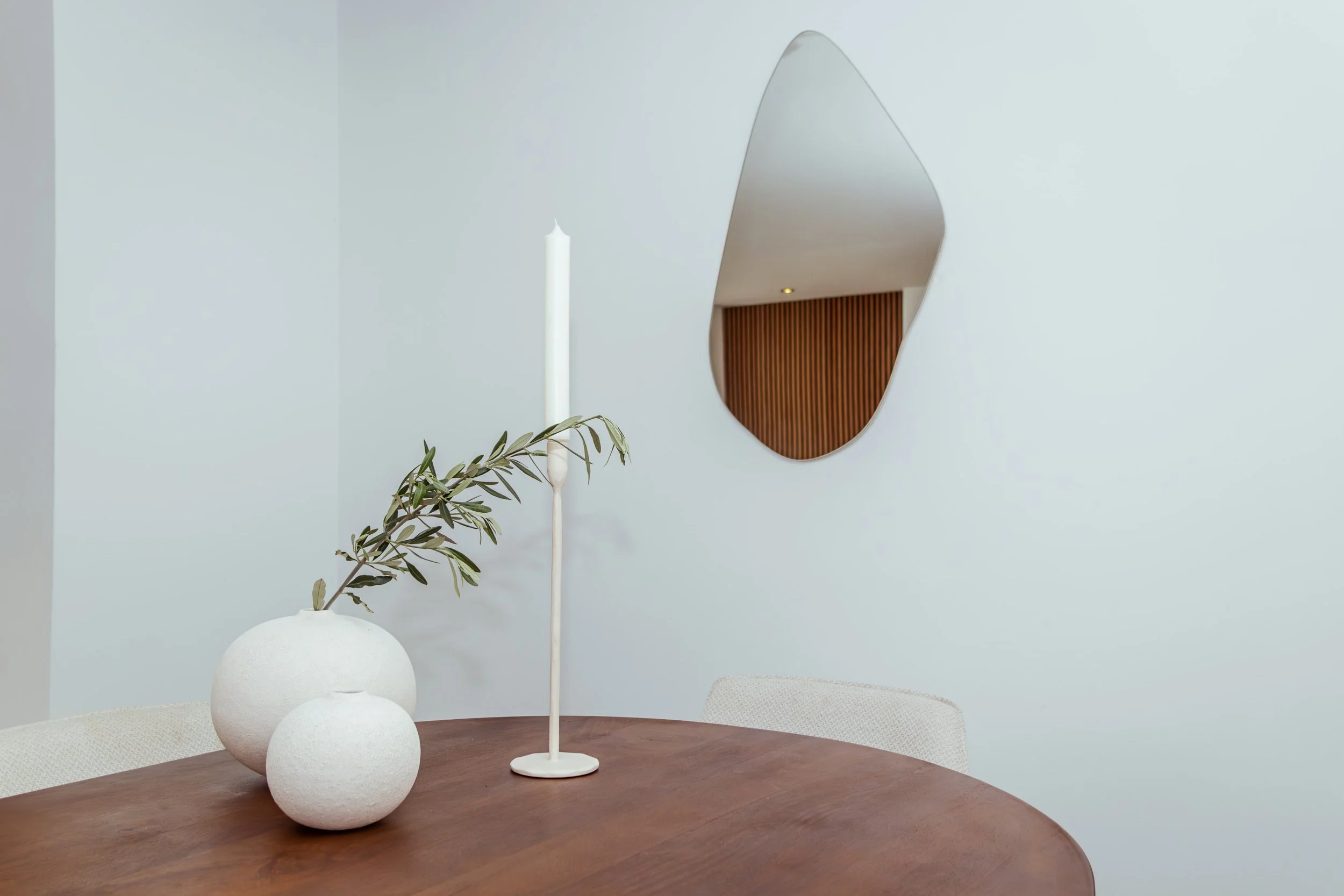 Een ronde houten tafel met witte vaas met groen takje, een witte kaars op een houder, en lichtkleurige stoelen, een spiegel aan de muur.