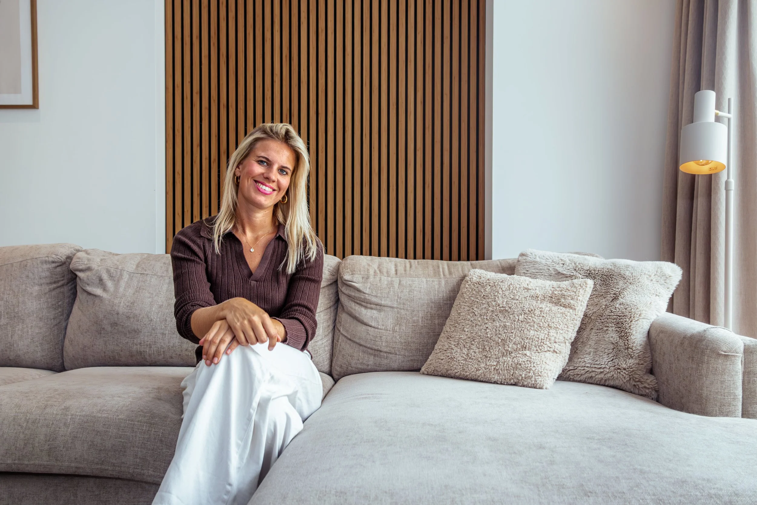 Vrouw zittend op een bank in een woonkamer, lachend en gekleed in een bruin shirt en witte broek, met een houten wand en een witte muur met een lamp op de achtergrond.