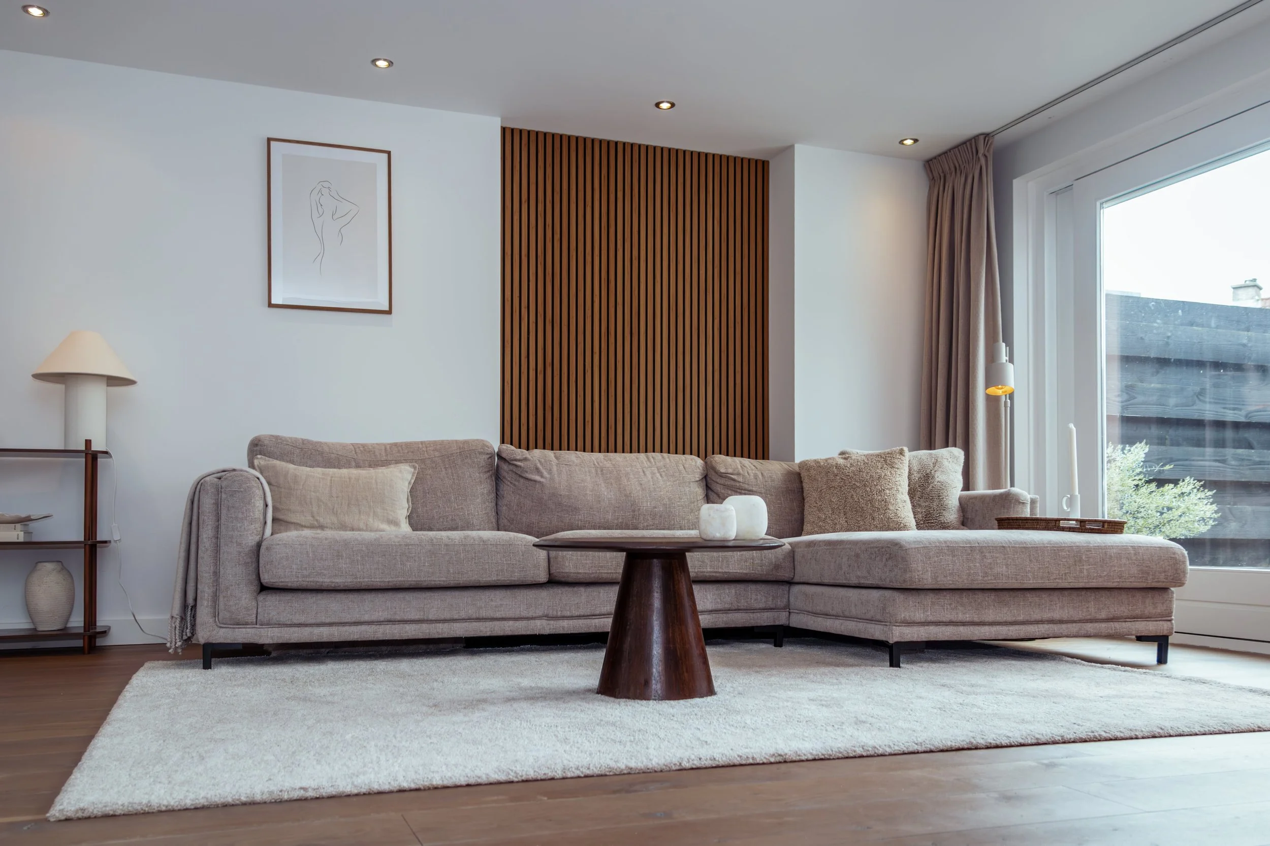 Een woonkamer met een lichtgrijze hoekbank, een houten salontafel met decoratieve objecten, een wit vloerkleed, een raam met beige gordijnen, een witte muur met een lijntekening en een badkamerlamp, en een houten panel op de muur.