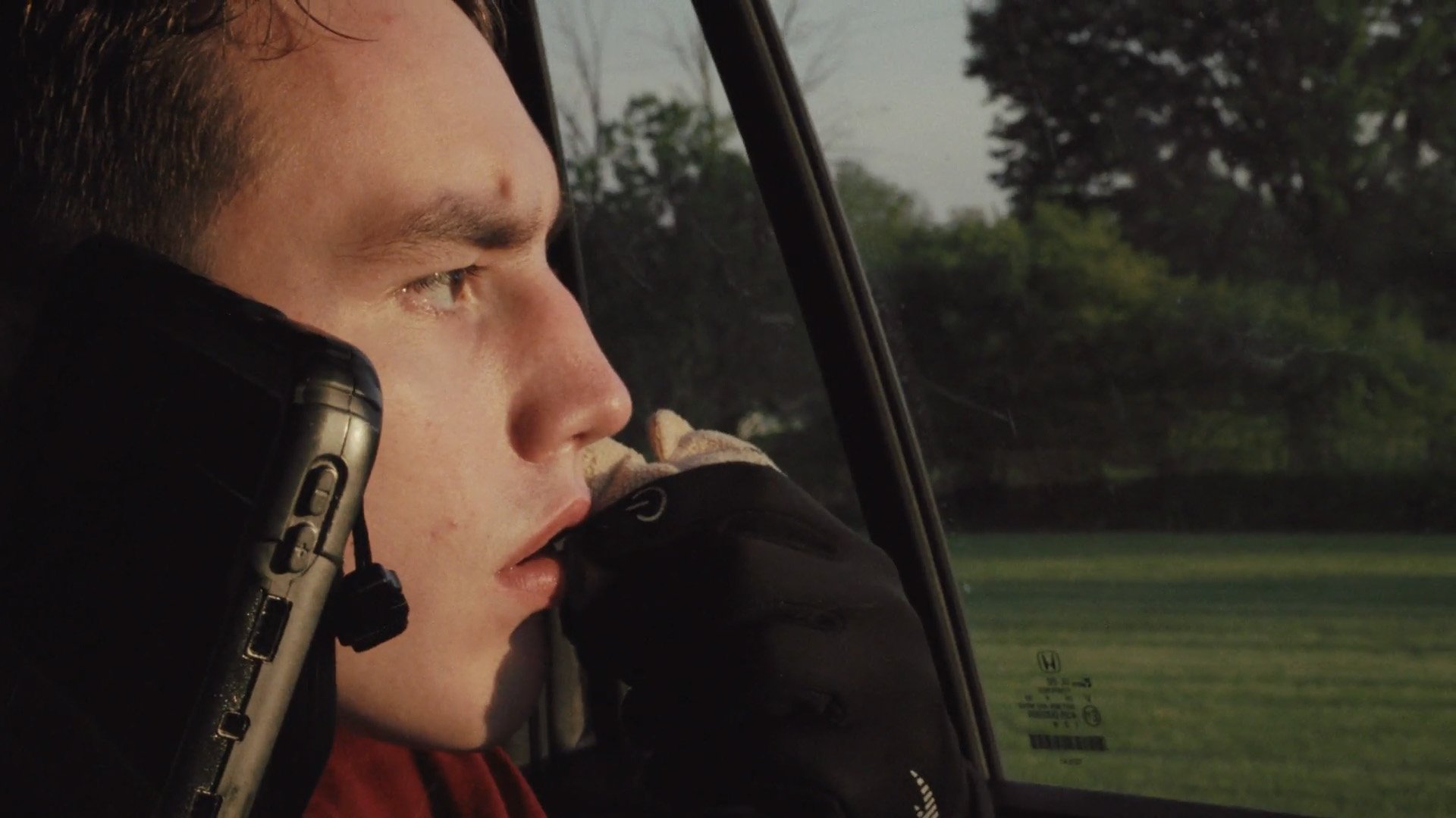 Close-up of a young man with short brown hair, seated in an aircraft, speaking on a device, with a serious expression, next to a window showing green trees and grass outside.