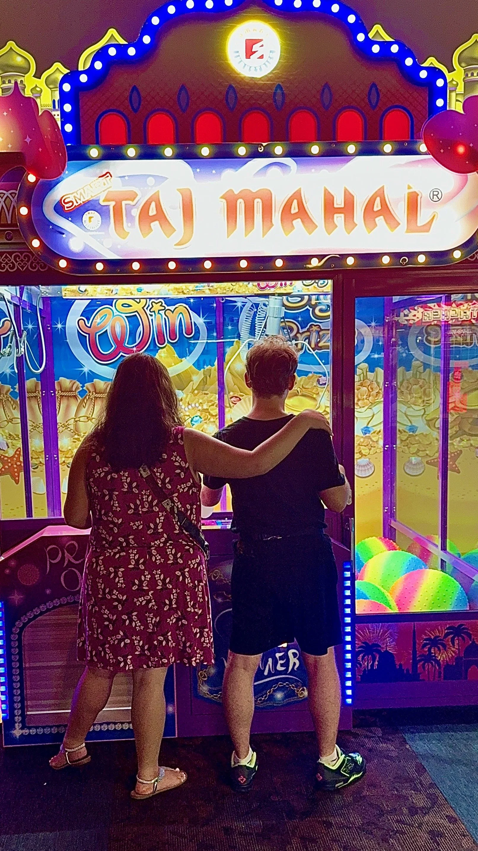 A man and woman are standing together at an arcade with a bright, colorful background of a casino. The woman has her arm around the man's shoulder, and they are facing the game machine, which is filled with arcade prizes like balls and stuffed toys. The sign above reads 'Taj Mahal' with colorful lights.