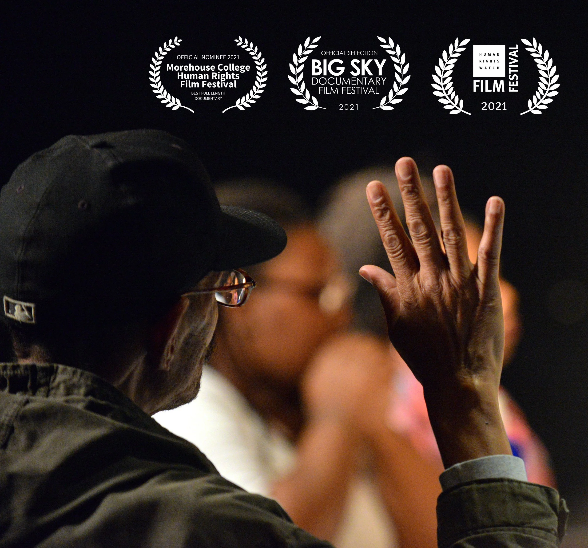 A person wearing a black cap and glasses raises their right hand during a Dorchester Clemente Course in the Humanities class, with others blurred in the background. Overlaid are three film festival laurels indicating awards from 2021.