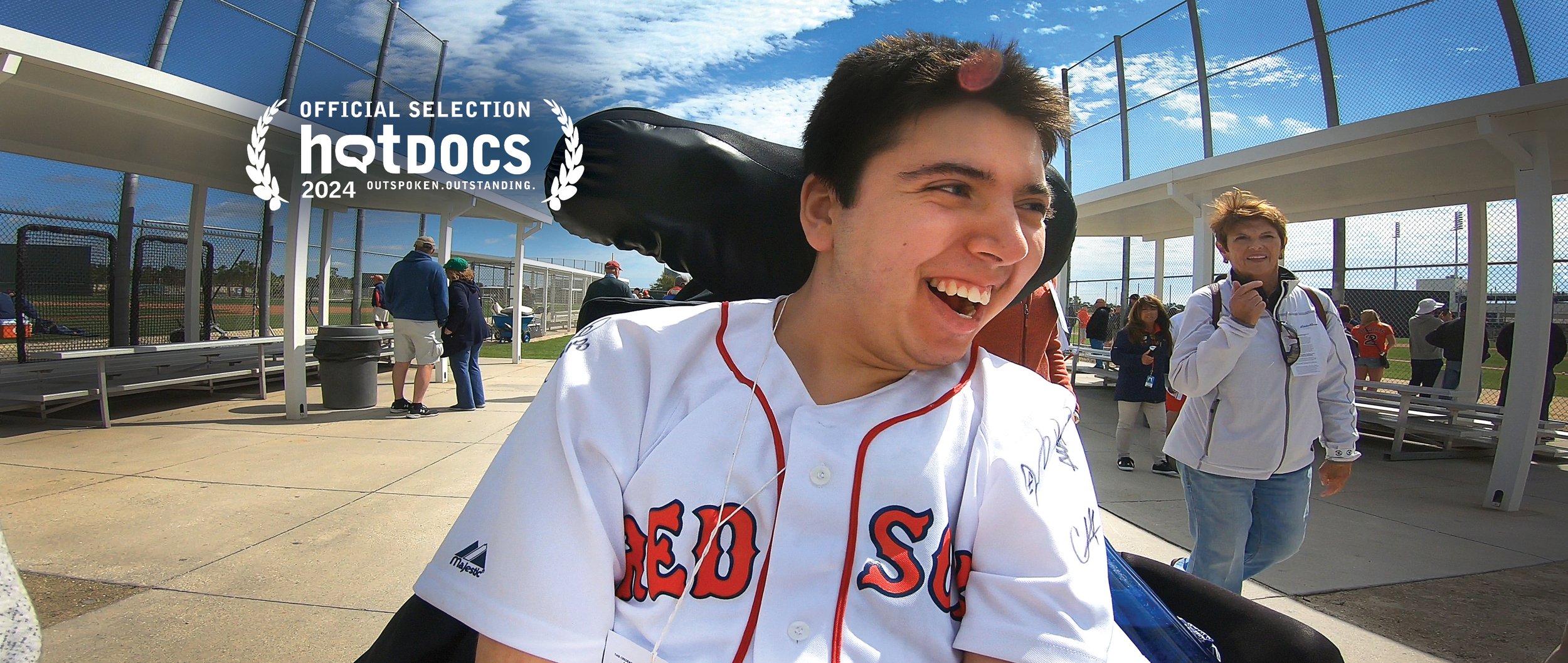 Samuel Habib smiling and wearing a Boston Red Sox jersey at a baseball field. People are in the background near the dugout and fenced field, under a cloudy sky.