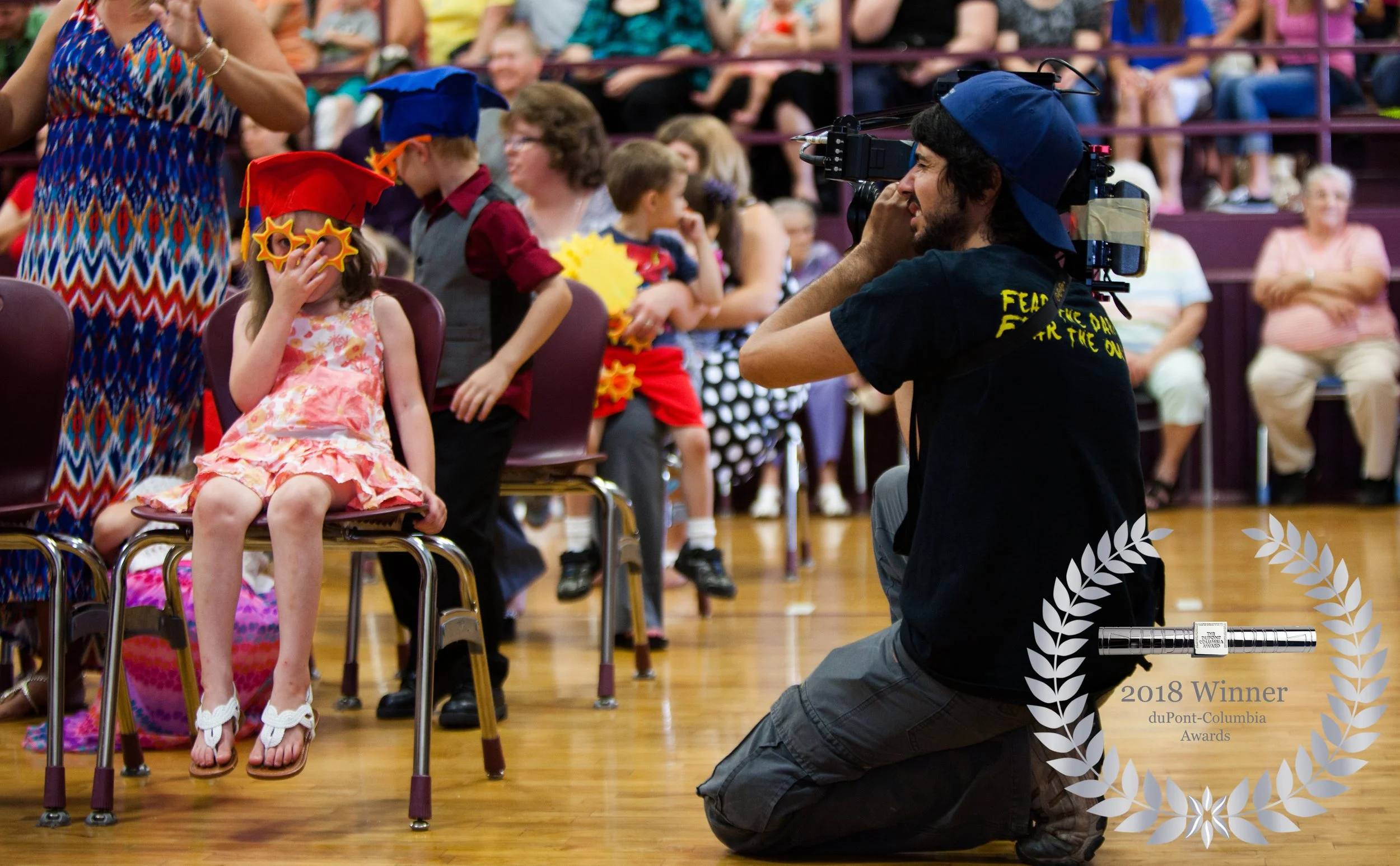 A young girl sitting on a chair at a Head Start graduation ceremony, wearing a red cap and star-shaped sunglasses, with her hand covering her face. Cinematographer Antonio Cisneros  is filming the event, kneeling on the floor.
