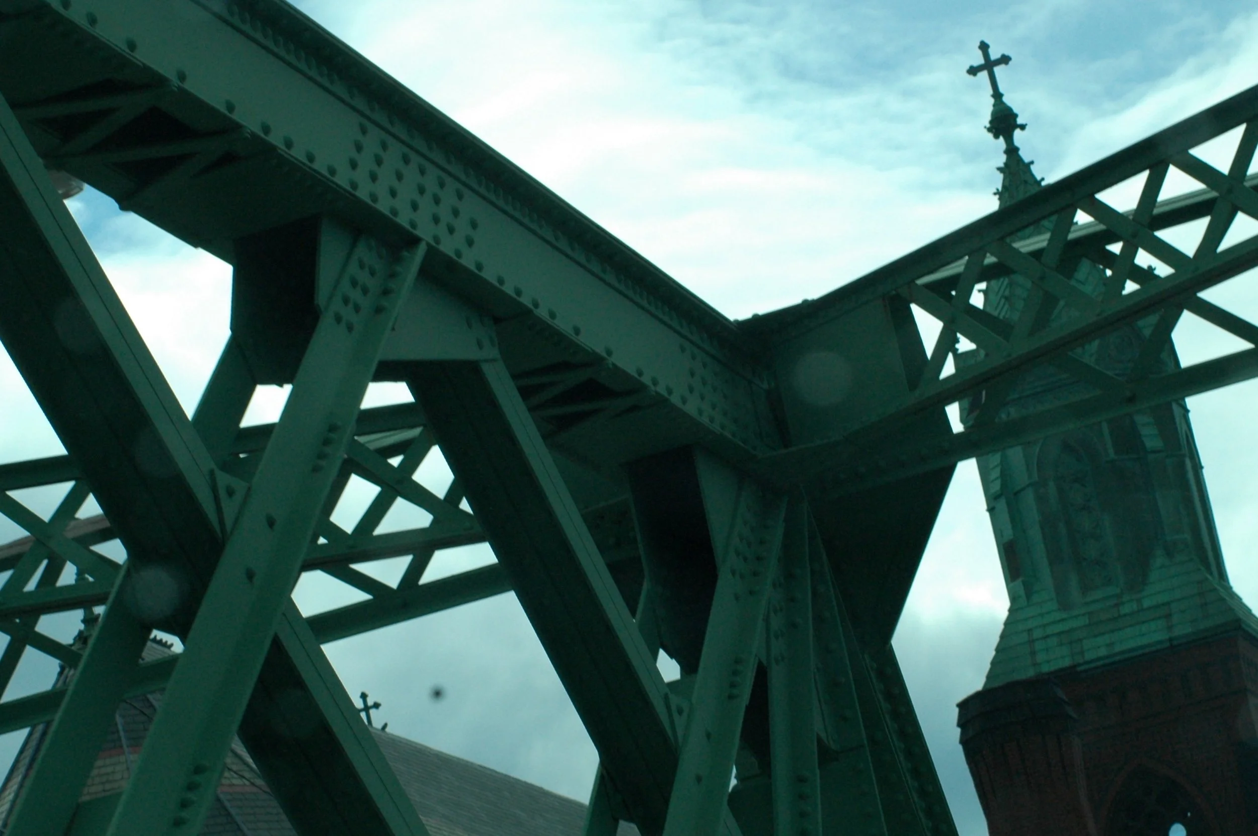 Low-angle view of a steel bridge structure with a church steeple with a cross in the background against a cloudy sky.