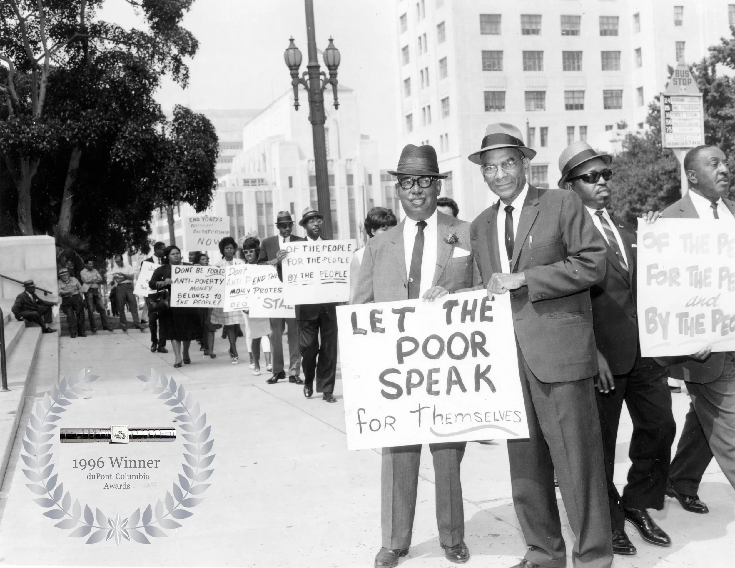 Black and white photo of a civil rights protest march with people holding signs and a group of men in suits and hats at the forefront. One sign reads 'Let the poor speak for themselves,' and other signs carry messages advocating against poverty and discrimination. There is a large building and trees in the background.