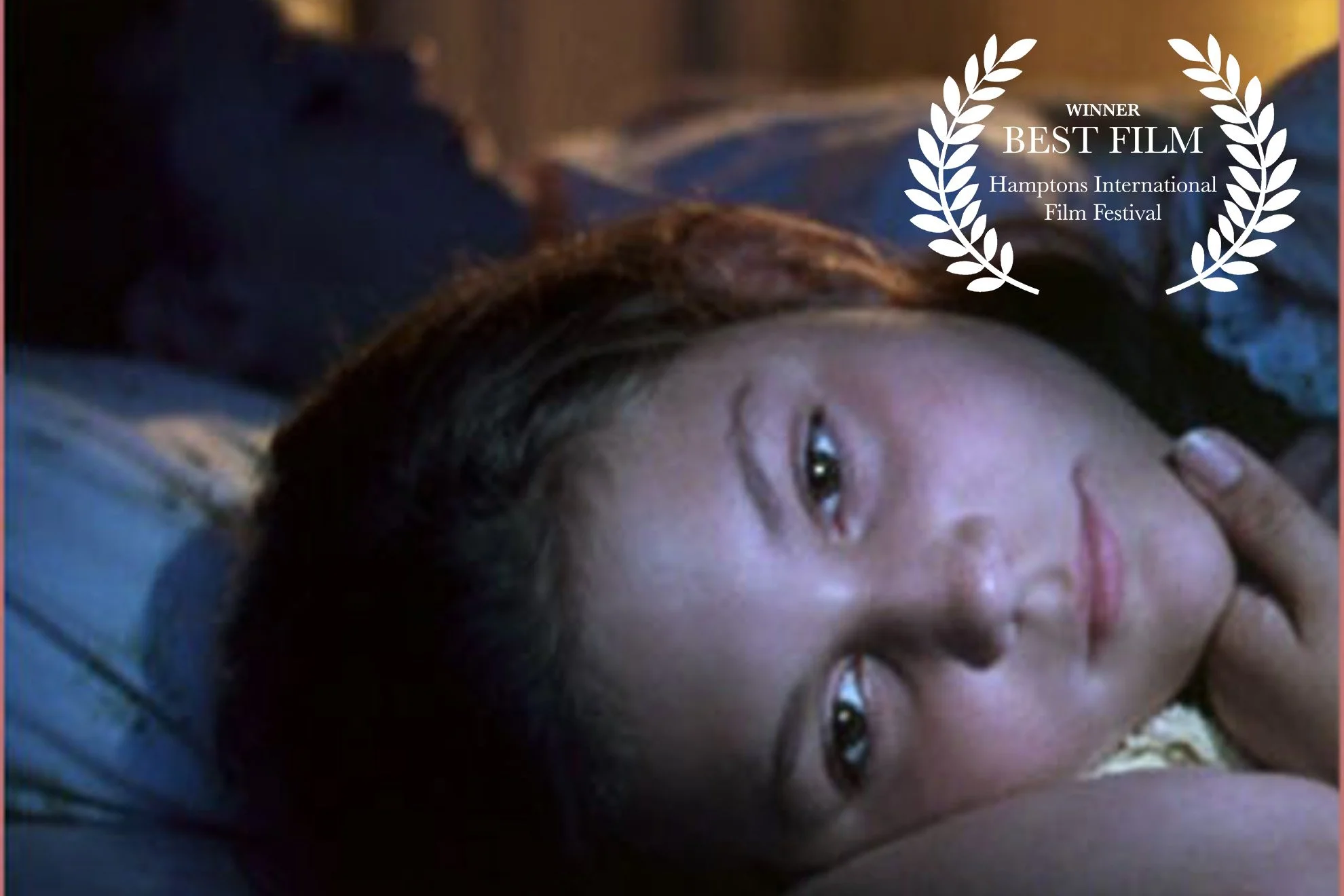 A woman lying on her side with her head resting on a pillow, looking directly at the camera with a gentle expression. There is a white film festival winner laurels graphic with text indicating 'Winner Best Film Hamptons International Film Festival' overlaying the upper right corner of the image.