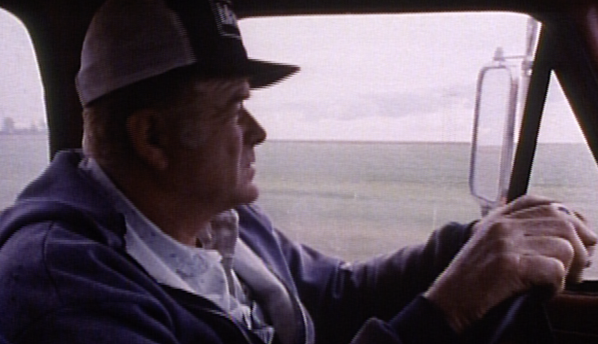 Farmer John Ferrari wearing a baseball cap driving a vehicle with a large side mirror, with a cloudy sky in the background.