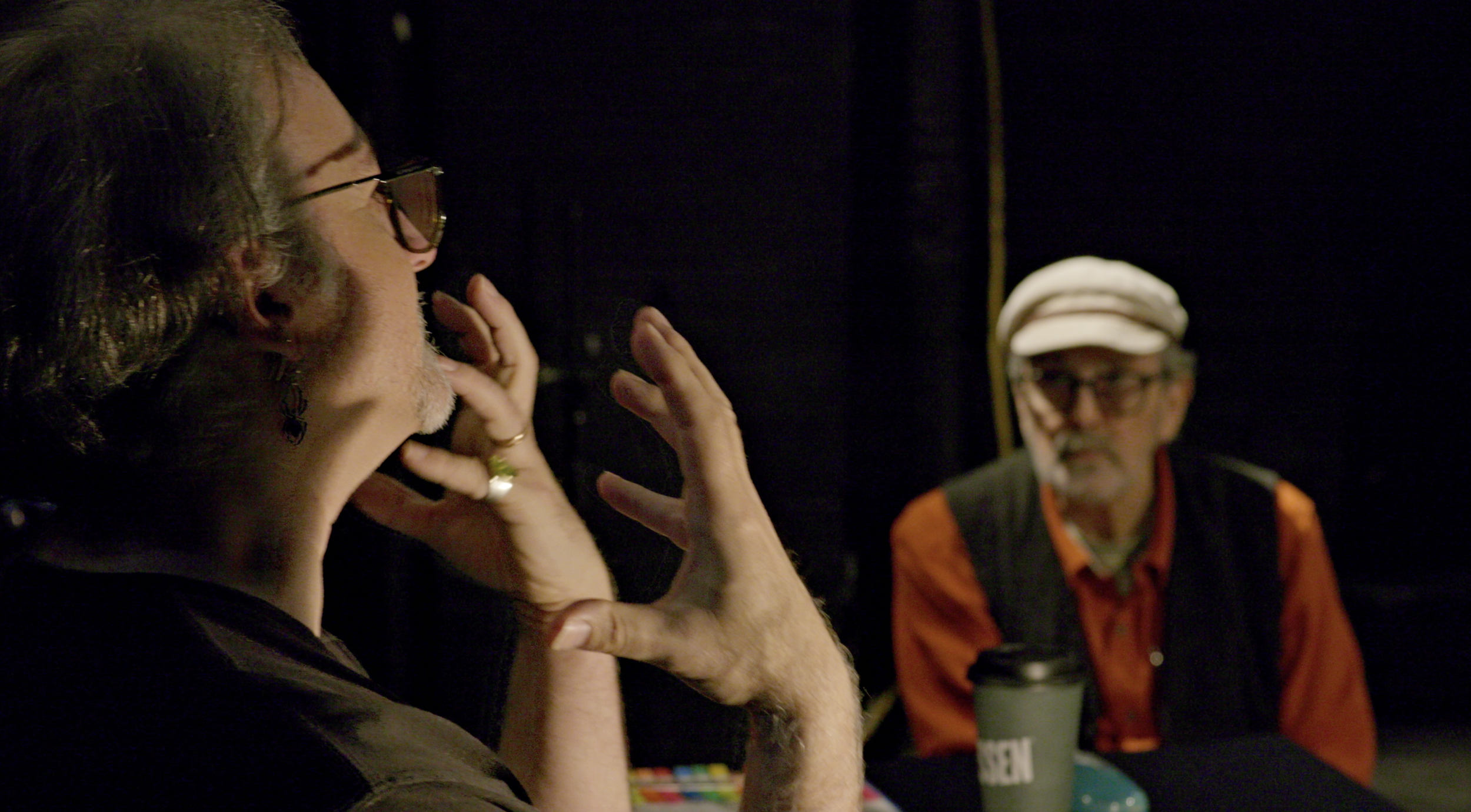 Two people with glasses and gray hair sitting at a table in a dark room, engaged in conversation. The one on the left is touching his chin and the one on the right is looking towards him, wearing a white cap and an orange shirt under a vest.