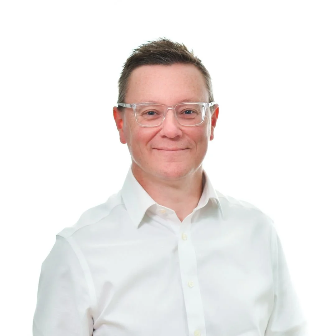 A man with short brown hair, wearing clear glasses and a white button-up shirt, smiling at the camera against a plain white background.