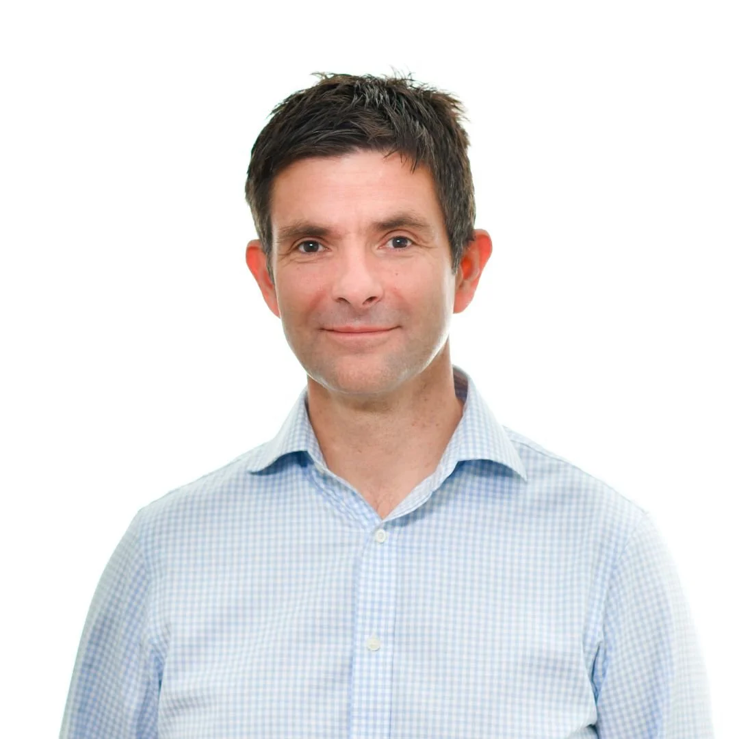Portrait of a man with short dark hair, wearing a light blue checkered button-up shirt, smiling softly, against a white background.