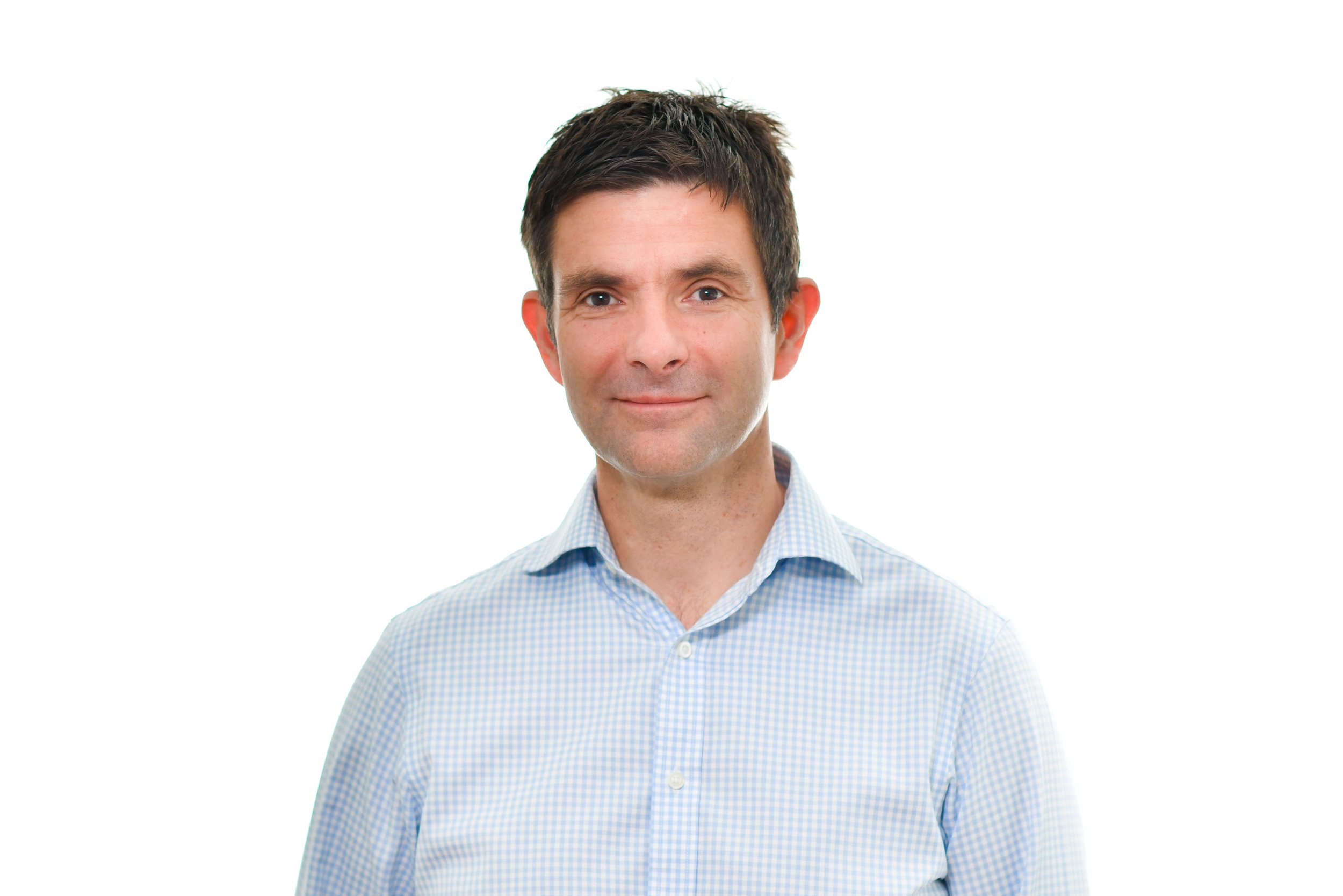 A man with dark hair and fair skin wearing a light blue and white checkered shirt, smiling softly against a white background.