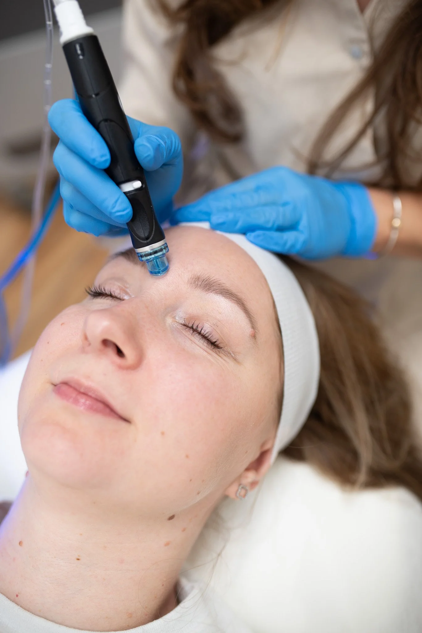 A woman receives a cosmetic facial treatment using a handheld device on her forehead while lying down with her eyes closed, wearing a white headband and a white shirt.