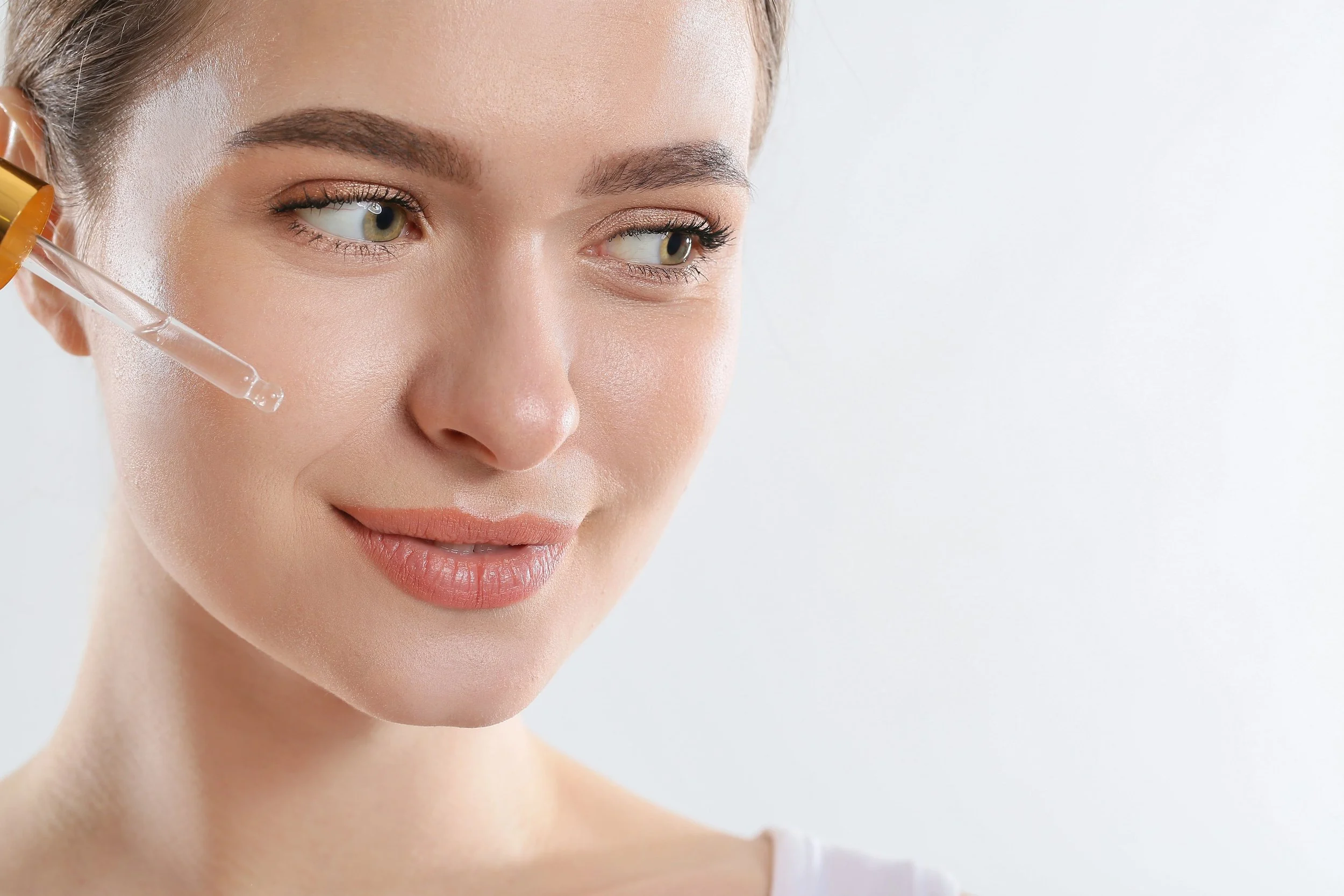 A woman with clear skin receiving a facial serum with a dropper from a bottle, against a plain white background.